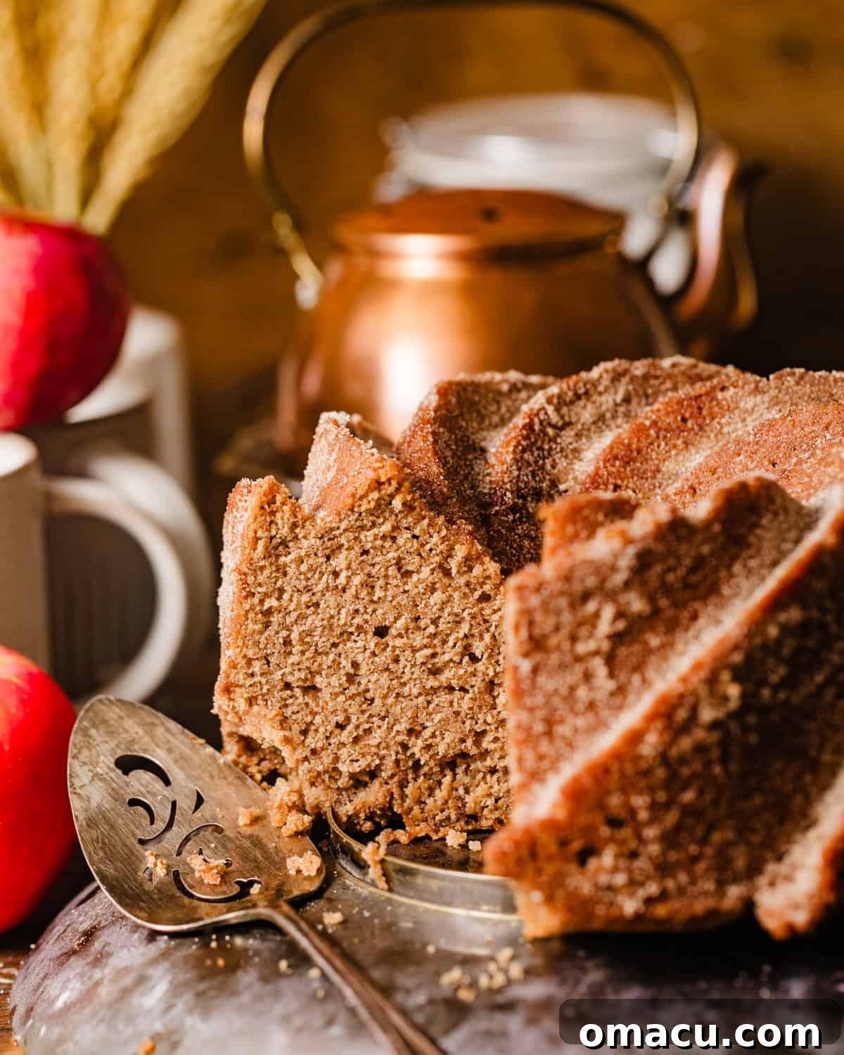 A whole Apple Cider Bundt Cake on an elegant cake stand, surrounded by fresh apples and a cake server, ready to be enjoyed.