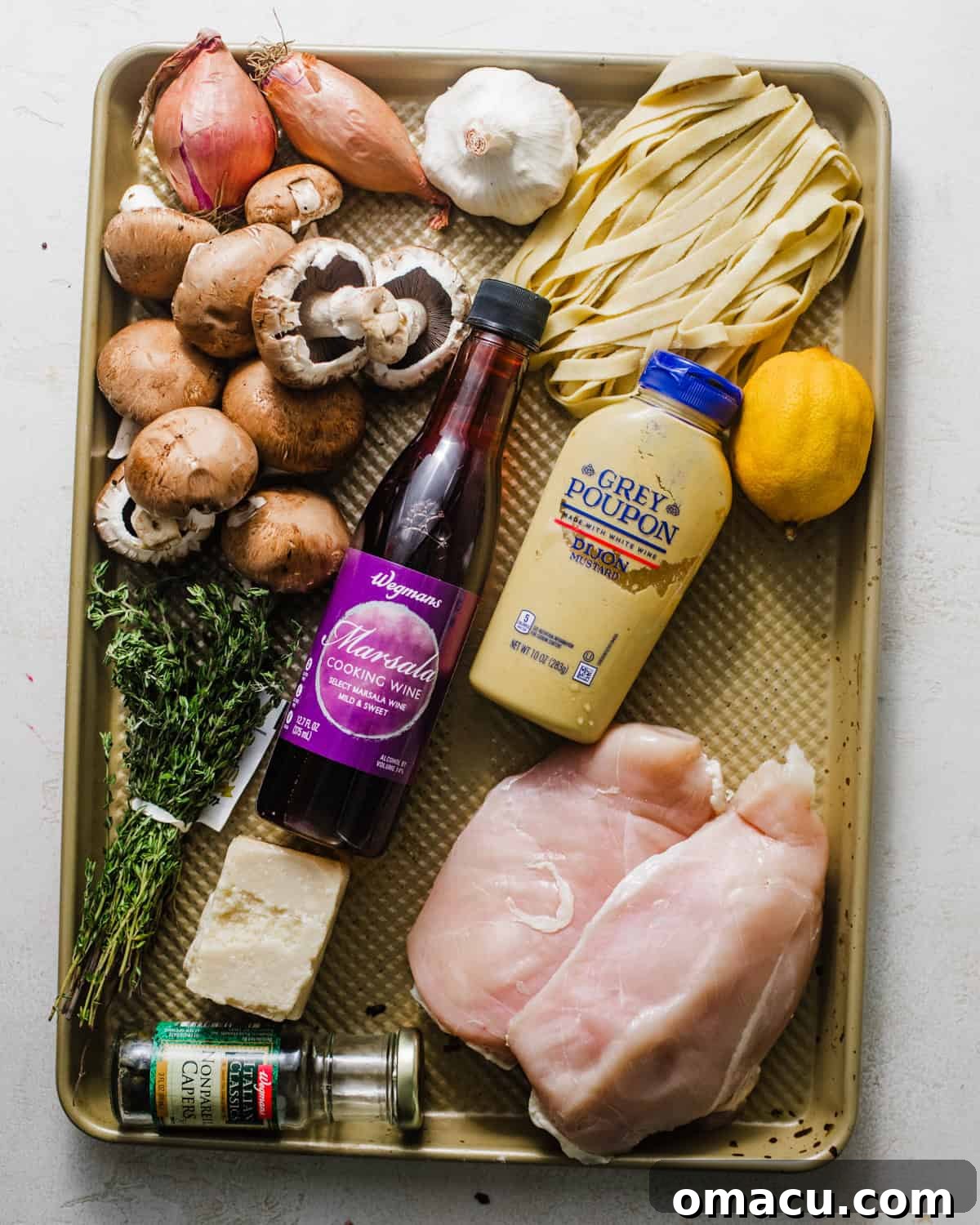 A collection of fresh ingredients laid out on a baking tray, including chicken breasts, shallots, mushrooms, capers, and herbs, ready for making chicken marsala fettuccine.