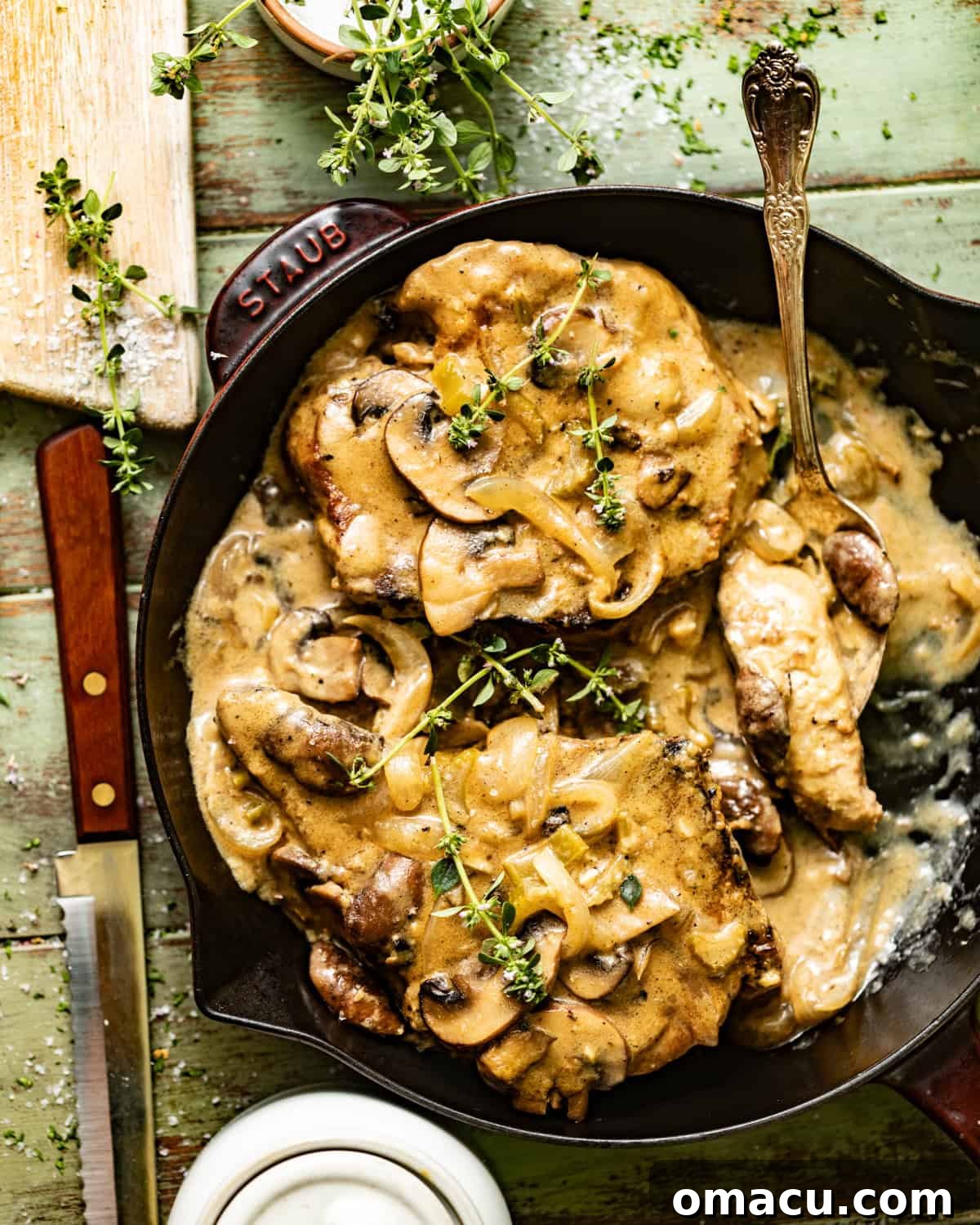 smothered pork chops in a skillet topped with fresh thyme in a mushroom and onion gravy. A close-up shot of the juicy pork chops immersed in the rich, brown gravy.