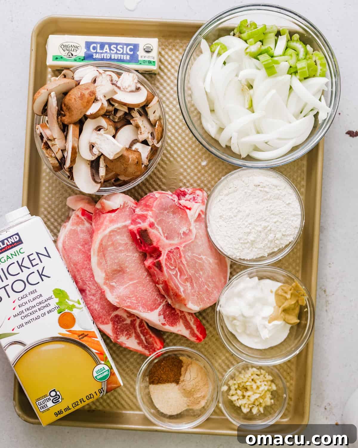 A collection of fresh ingredients laid out on a baking sheet, ready for smothered pork chops. Includes bone-in pork chops, sliced onions, celery, garlic cloves, fresh mushrooms, a variety of seasonings, all-purpose flour, Dijon mustard, chicken broth, heavy cream, and butter.