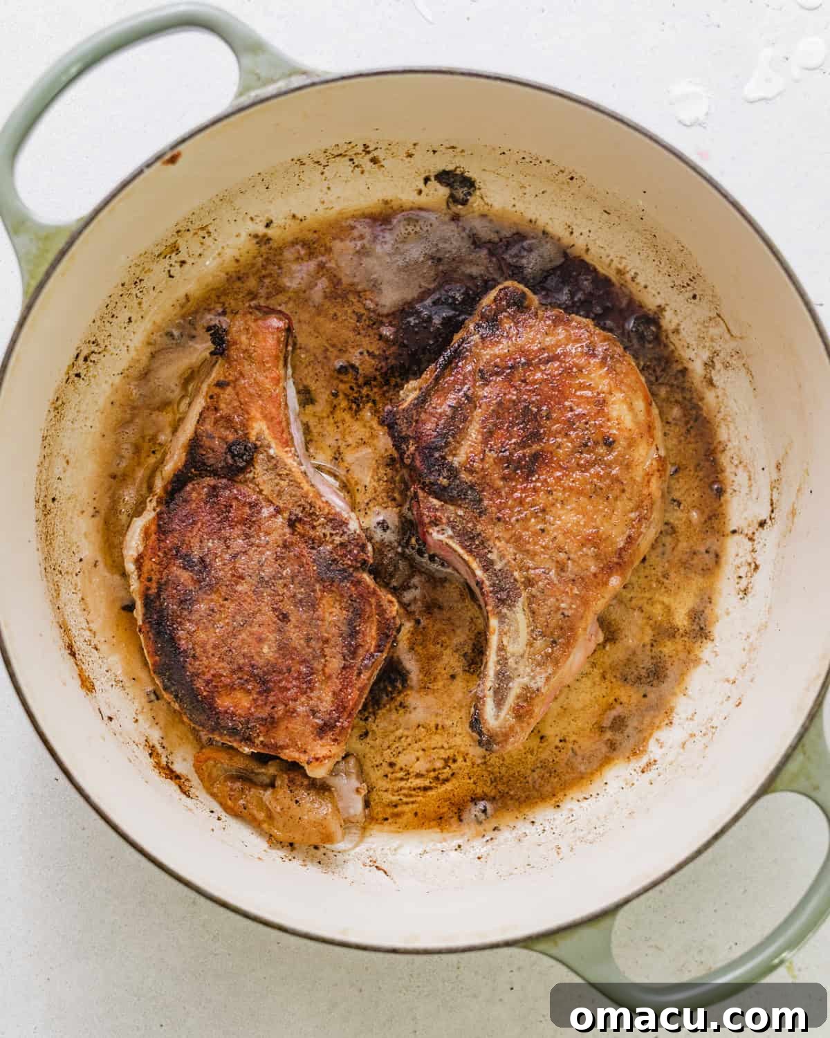 Two golden-brown, seared pork chops cooking in a large, deep skillet.