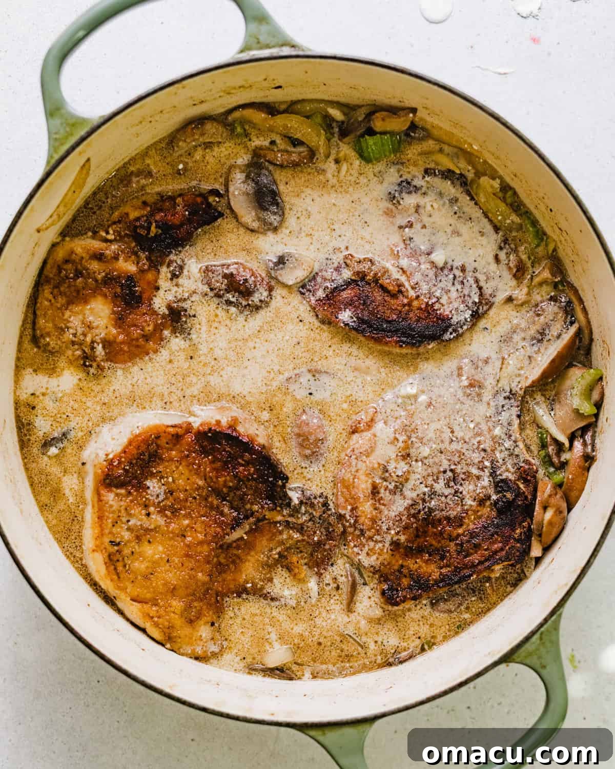 Pork chops simmering in a rich, creamy gravy with mushrooms and onions, topped with fresh thyme, in a deep skillet.