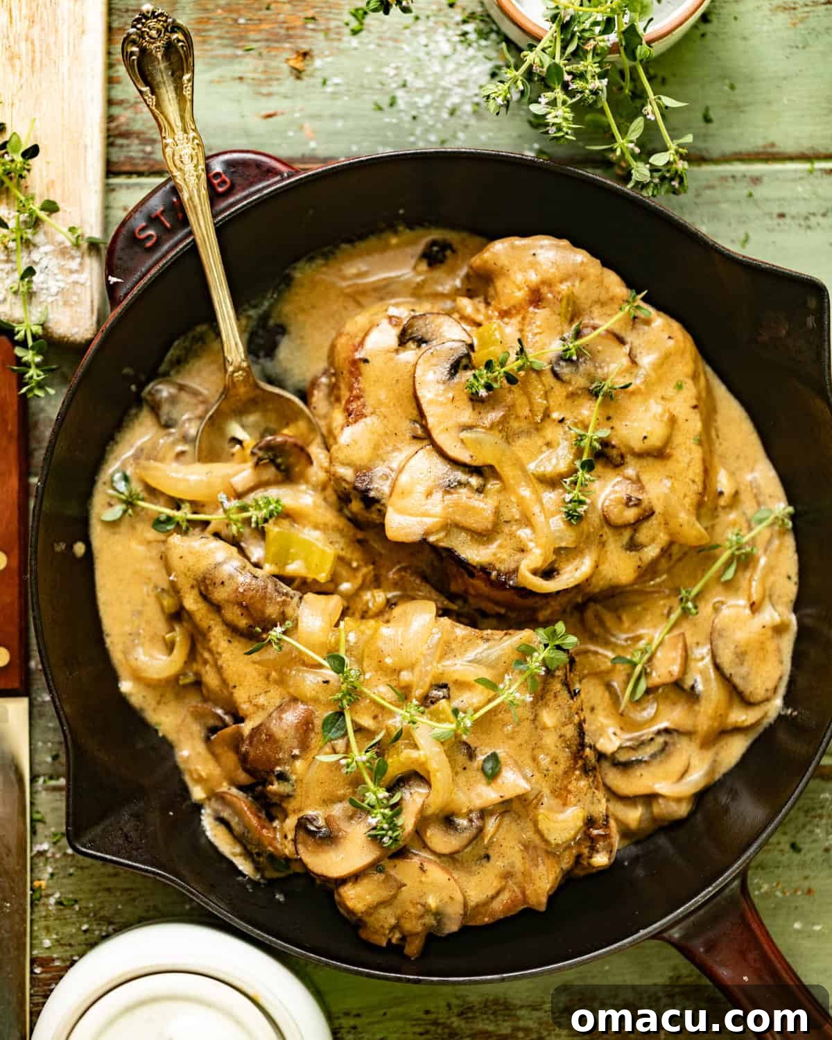 A close-up of a tender smothered pork chop in a skillet, partially covered by the creamy mushroom and onion gravy. A serving spoon rests in the pan, ready to dish out.