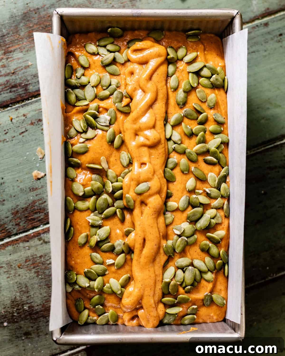 Smooth, vibrant orange-hued pumpkin bread batter is carefully poured into a parchment-lined loaf pan, then topped with a generous, even sprinkle of raw pumpkin seeds, creating a visually appealing crust before baking.