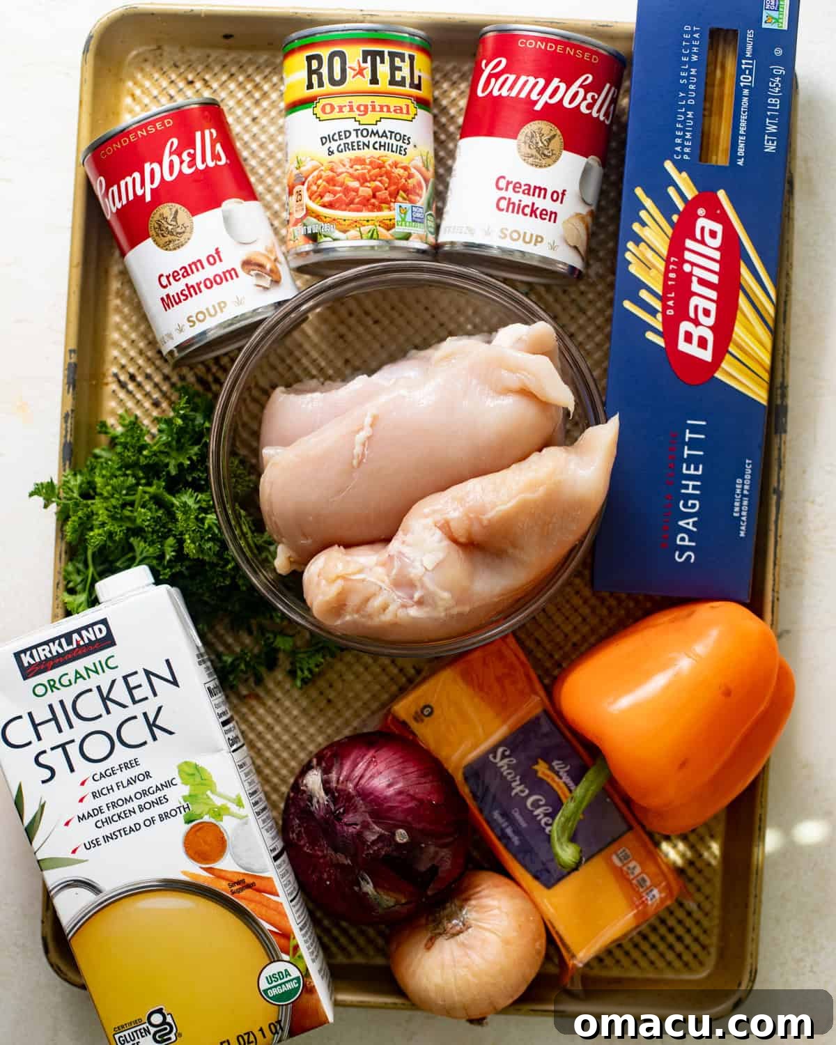 ingredients laid out on a baking tray for slow cooker chicken spaghetti - including boneless skinless chicken breasts, chicken stock, bell peppers, onions, parsley, cream of chicken soup, cream of mushroom soup, Rotel diced tomatoes, sharp cheddar cheese, garlic, and spaghetti noodles.