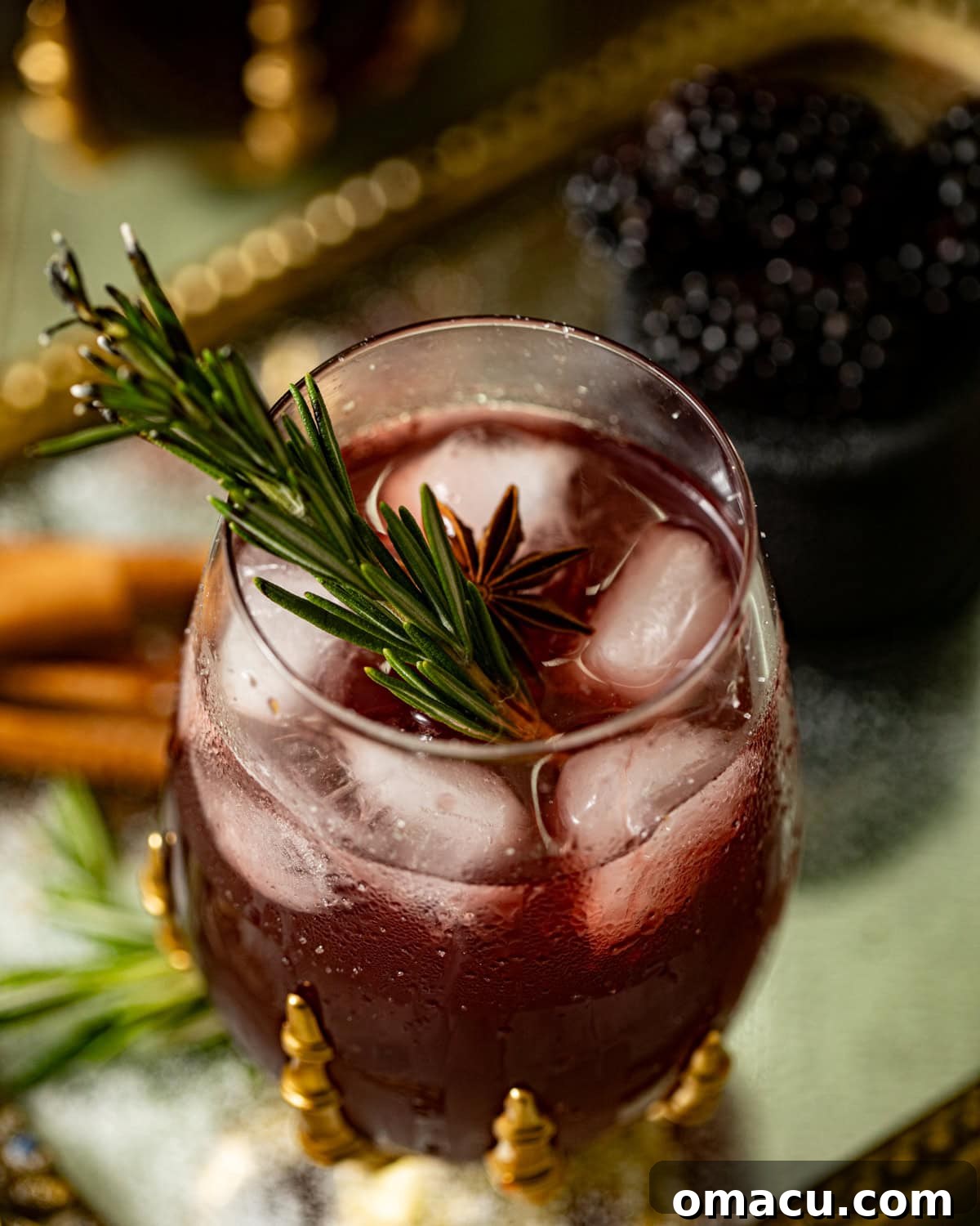 A selection of ingredients for the Witches Brew cocktail, including fresh blackberries, a bottle of bourbon, apple cider, and a star anise pod, artfully arranged on a reflective golden surface.