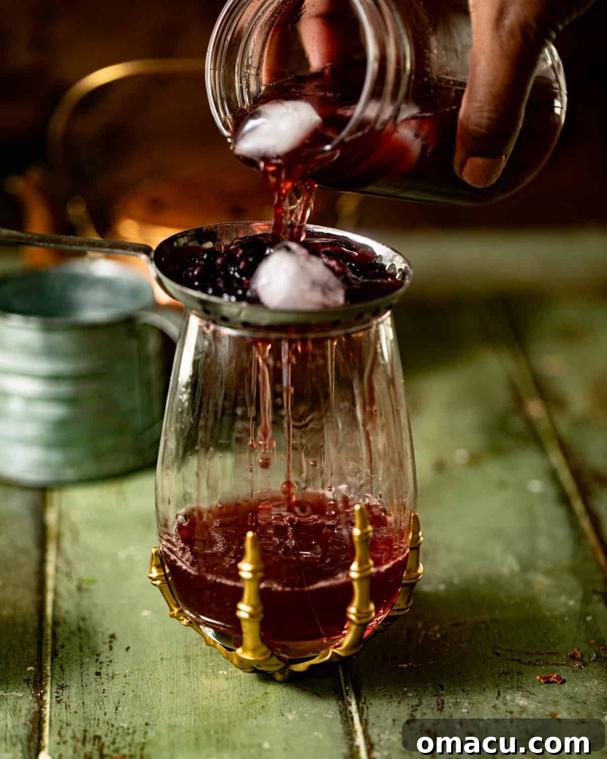 The prepared Witches Brew cocktail mixture being strained into a tall highball glass filled with ice, revealing a smooth, rich liquid.