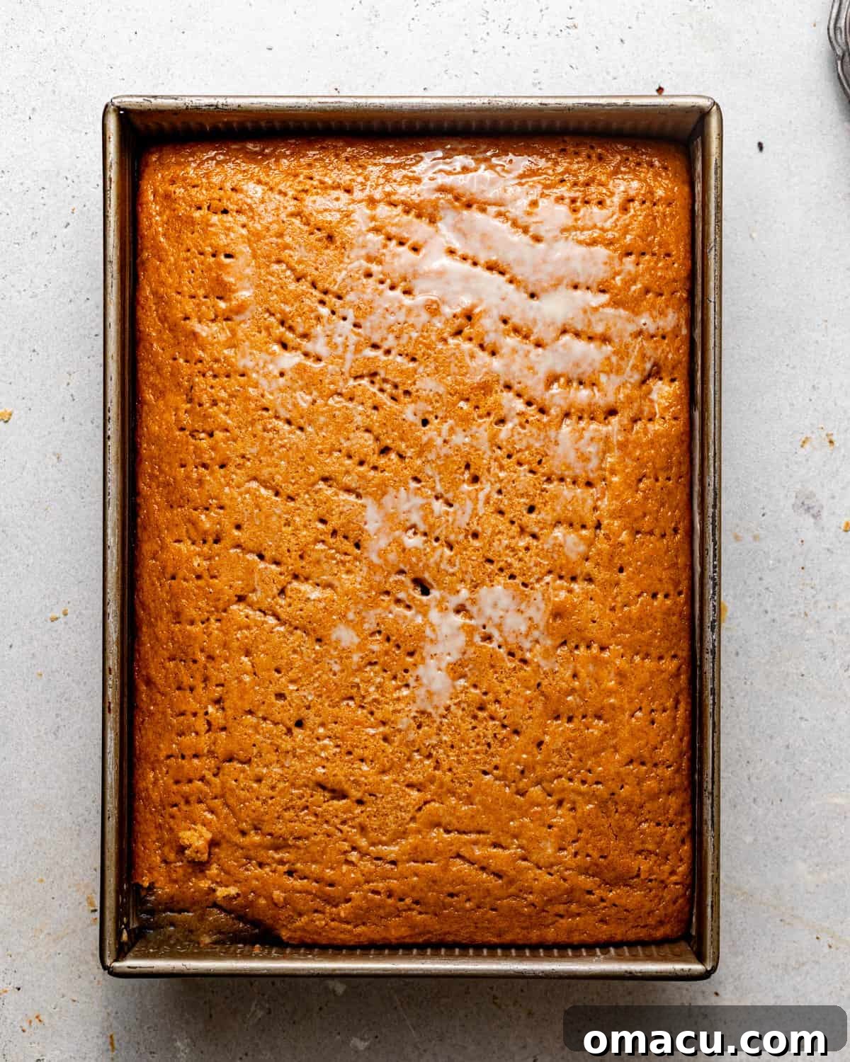 A freshly baked tres leches cake in a baking dish, generously poked with fork holes, with the three-milk mixture being poured over the top to soak in.