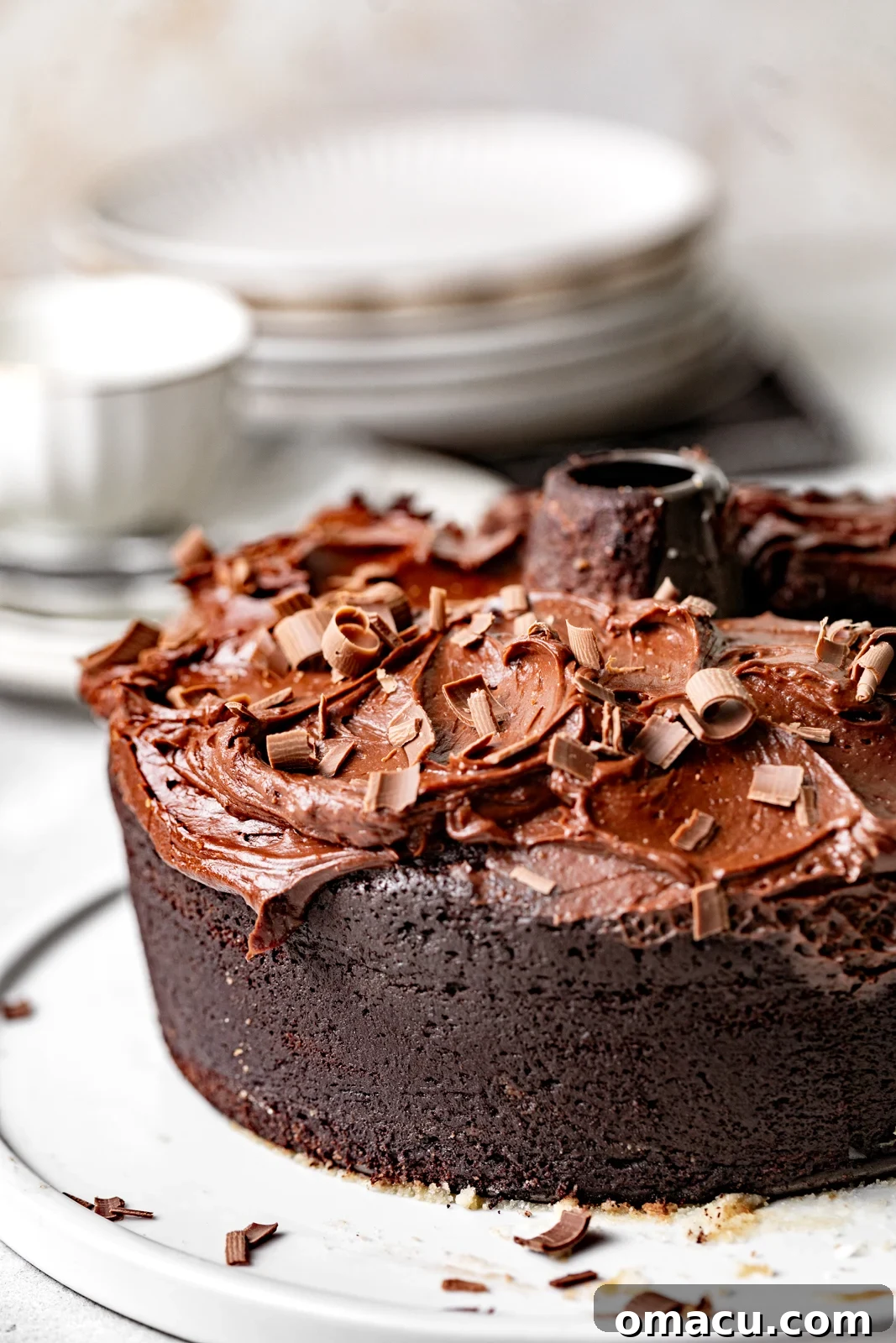 A freshly baked chocolate bundt cake rests elegantly on a cake plate, surrounded by smaller serving plates and artfully shaved chocolate in the background, hinting at a decadent dessert experience.