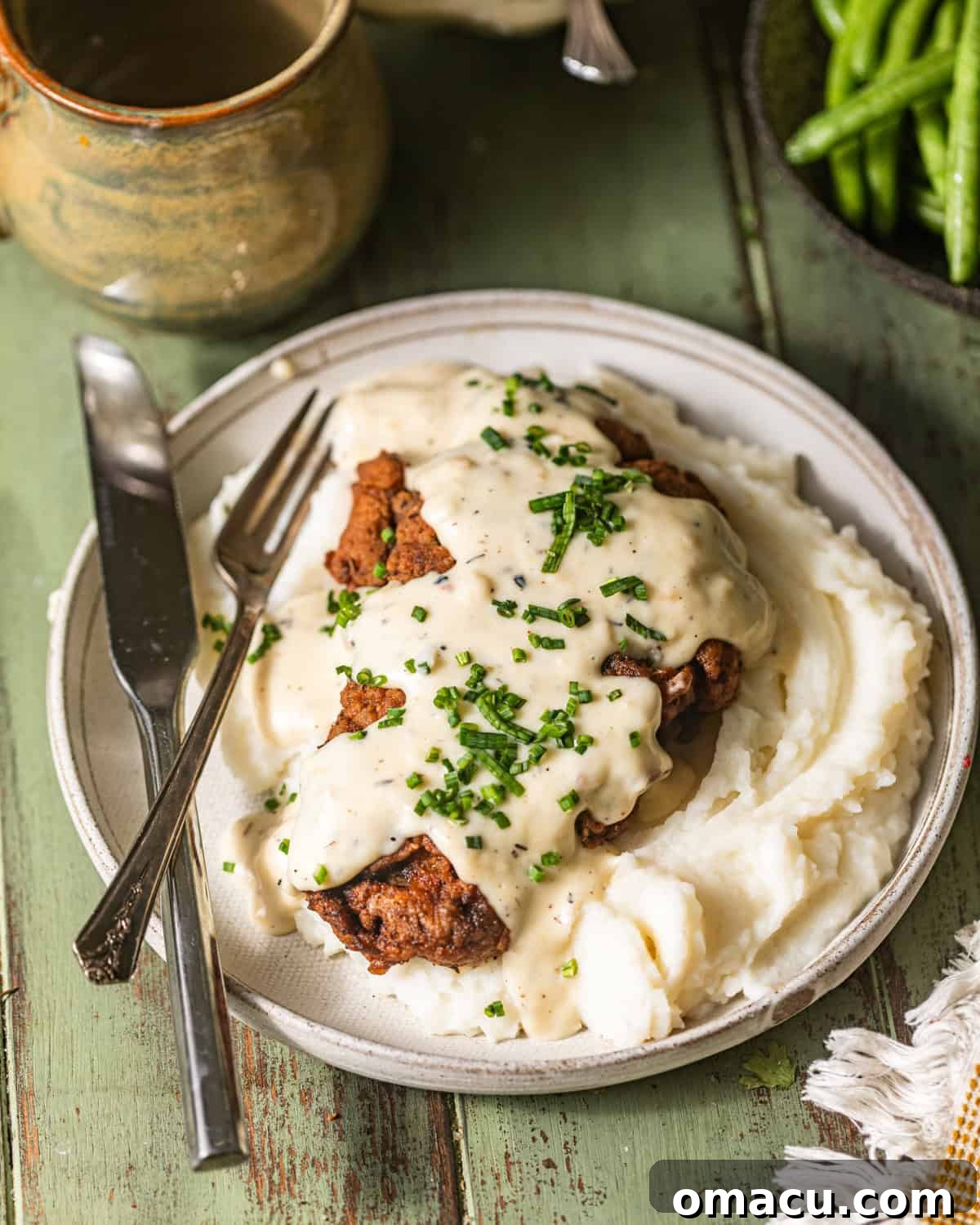 A plated chicken fried steak, garnished with fresh chives and generously covered in creamy white gravy, with a fork and knife ready for dining.