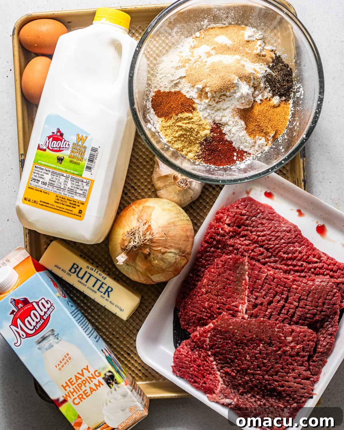 A flat lay photograph showcasing the essential ingredients for making chicken fried steak: cube steaks, fresh buttermilk, garlic, onion, all-purpose flour, a selection of robust seasonings, butter, cooking oil, farm-fresh eggs, rich heavy cream, and whole milk.