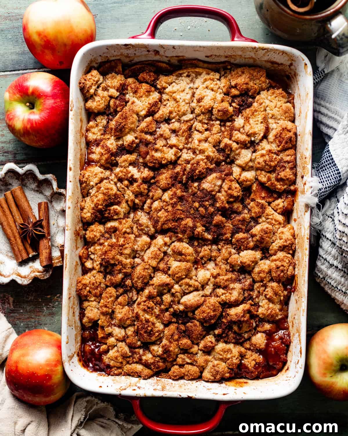 Overhead photo of a golden-brown apple cobbler in a baking dish, surrounded by fresh apples and cinnamon sticks, highlighting its rustic appeal.