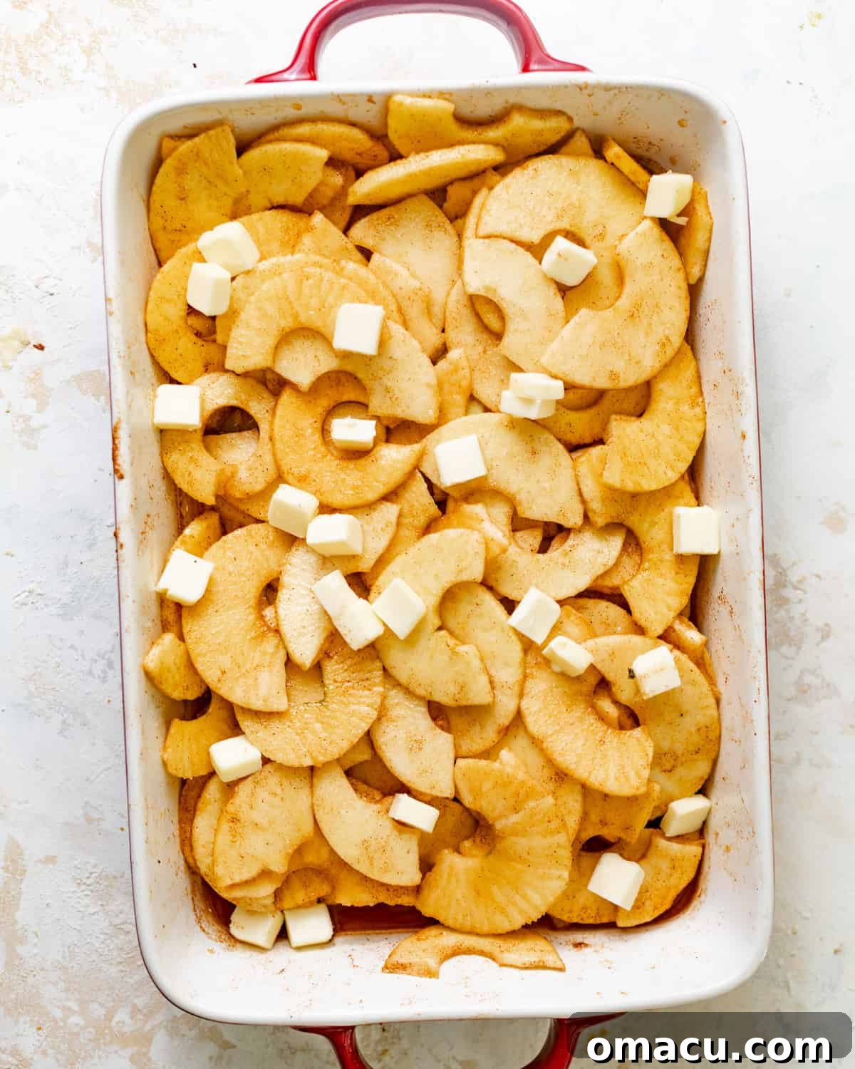 The apple cobbler filling is smoothed into an even layer in the baking dish, topped with small cubes of cold butter.