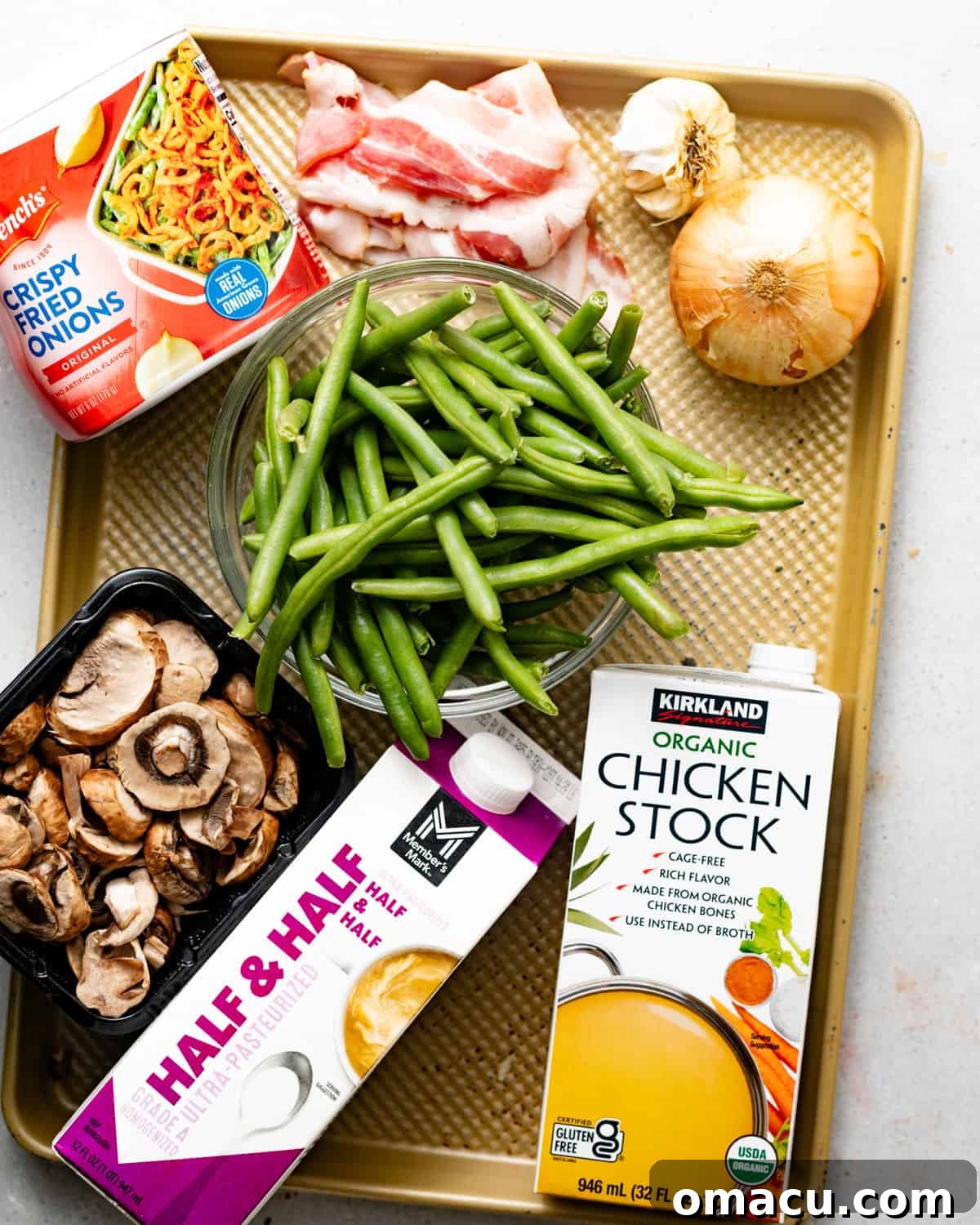 A beautifully arranged selection of fresh ingredients for green bean casserole, including vibrant green beans, sliced onions and mushrooms, half and half, a variety of spices, chicken bouillon paste, crispy bacon, French fried onions, garlic, chicken broth, and Dijon mustard.