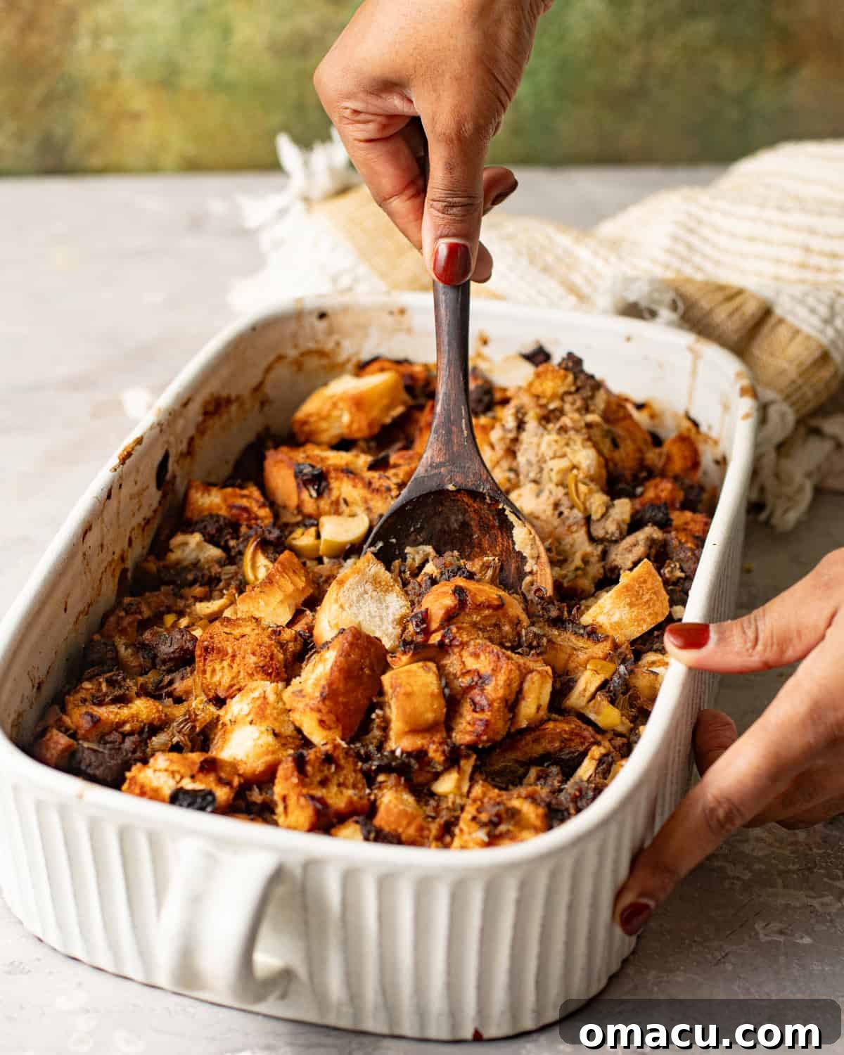 A wooden spoon scooping out golden brown sage and sausage stuffing from a baking dish.