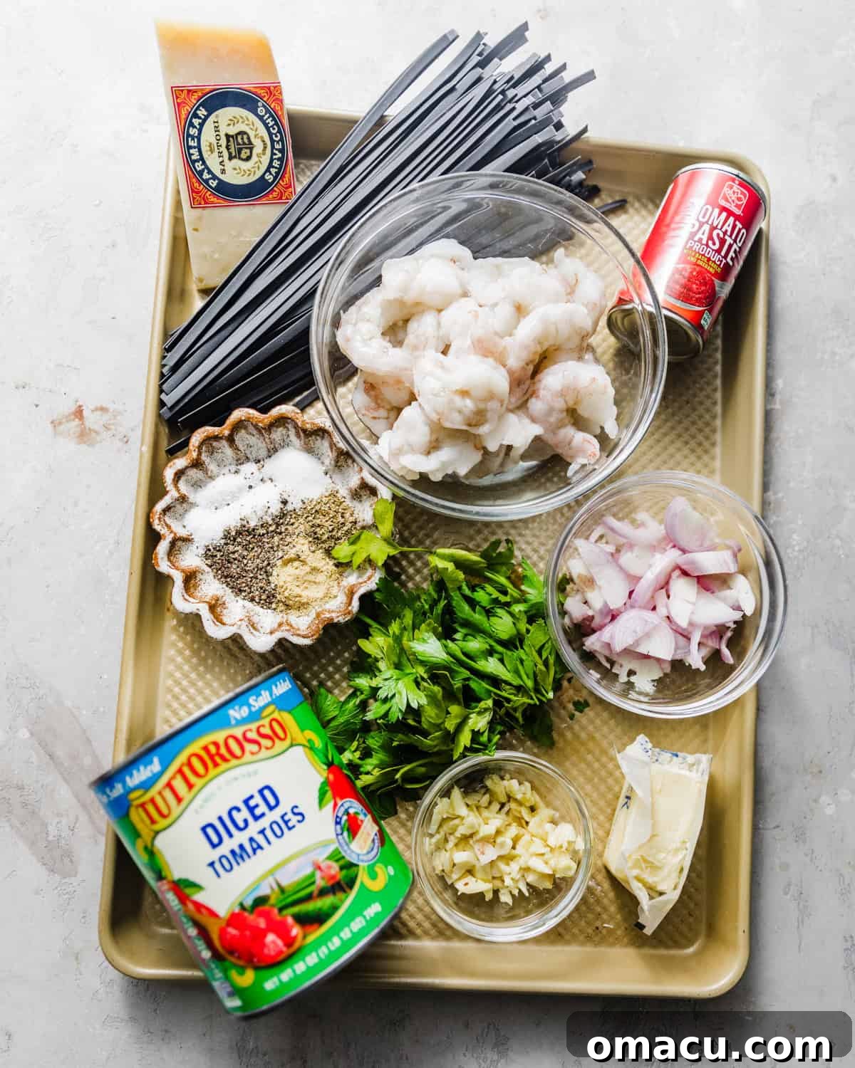 A baking tray displays all the essential ingredients for squid ink pasta with seafood, including uncooked black pasta, fresh shallots, garlic cloves, vibrant parsley, a can of crushed tomatoes, tomato paste, a bottle of white wine, grated Parmesan cheese, raw shrimp, fresh scallops, mussels, and various seasonings.