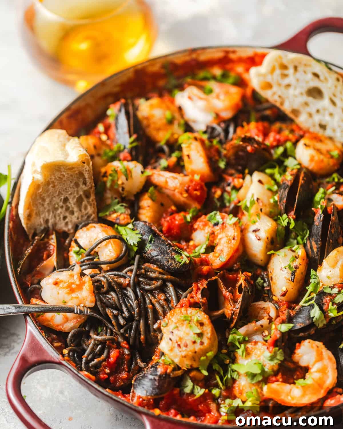 A large skillet filled with dark squid ink pasta, shrimp, mussels, and scallops, tossed in a rich red sauce, with a piece of crusty bread nearby.