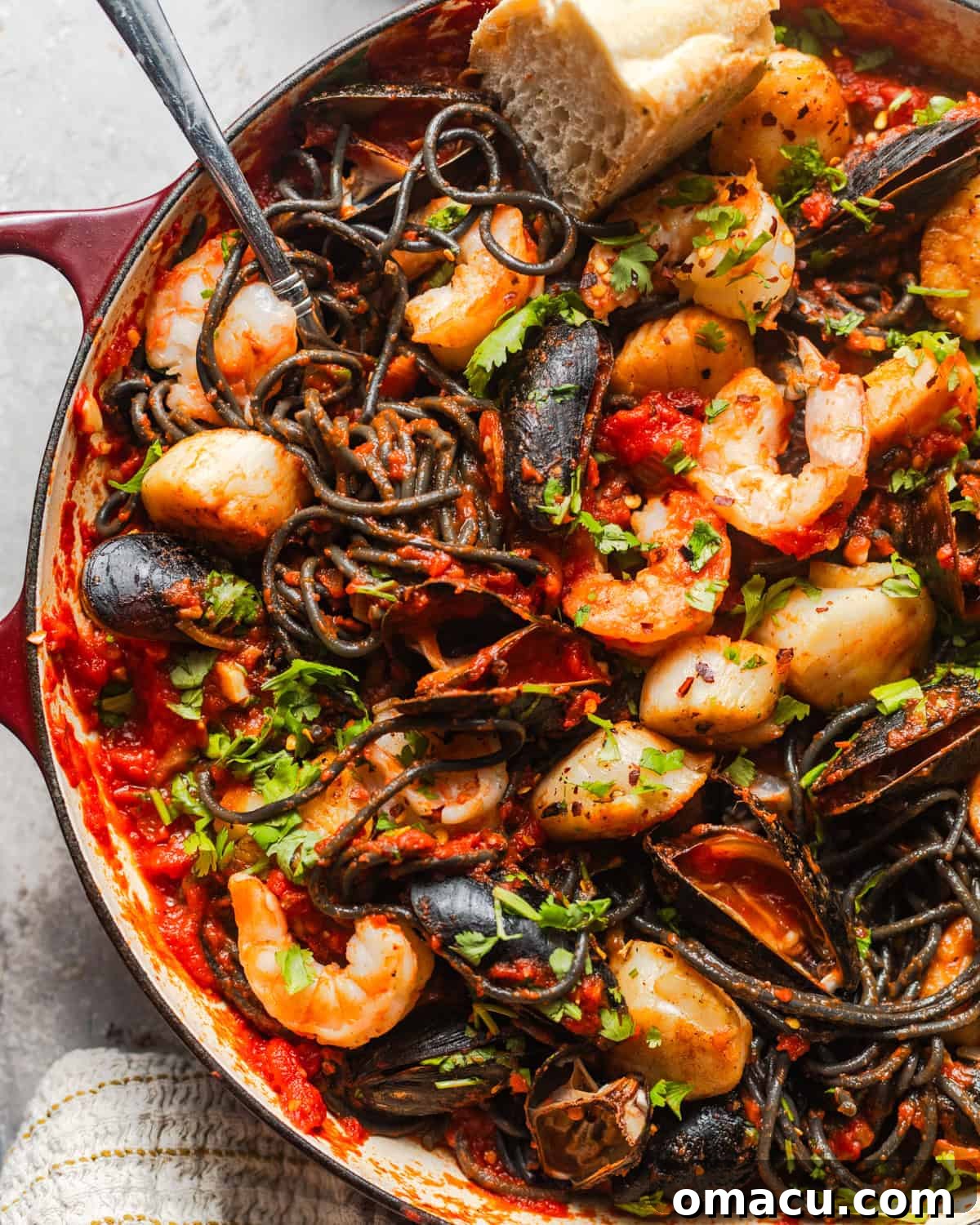An intimate close-up, overhead view of the dark squid ink pasta generously coated in sauce, with various seafood pieces visible in a skillet, ready to be served.