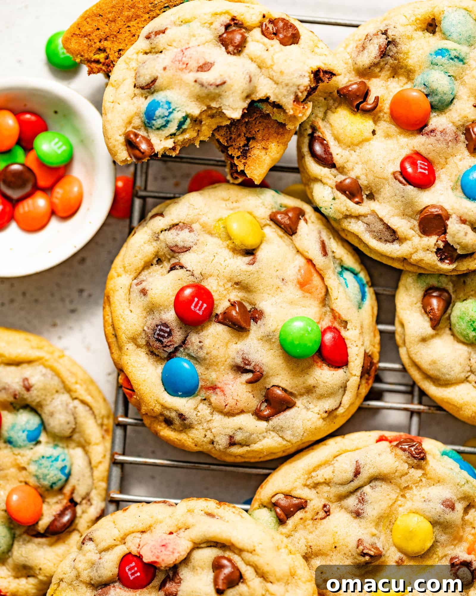 Freshly baked M&M chocolate chip cookies cooling on a wire rack, with a bowl of colorful M&M candies in the background.