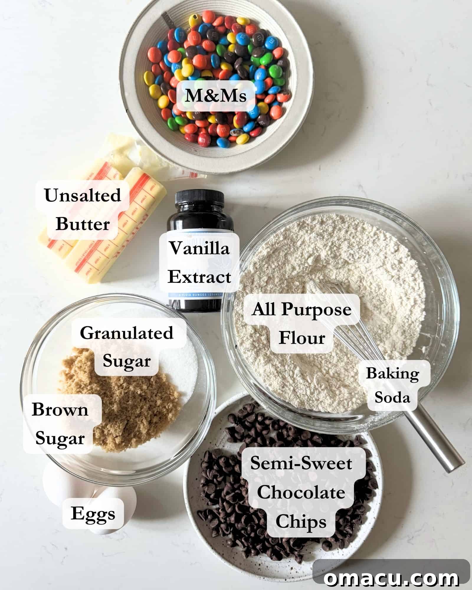 A curated display of all the essential ingredients for baking chocolate chip cookies with M&Ms: flour, sugar, brown sugar, butter, salt, baking soda, colorful M&M candies, chocolate chips, vanilla bean paste, eggs, and ground nutmeg.