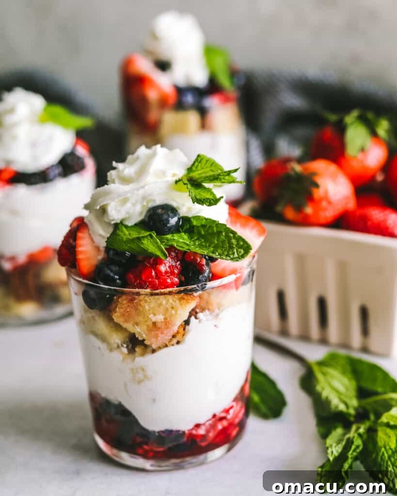 Mixed Berry Trifles in clear glass cups, garnished with mint, with more fresh berries scattered around for decoration.