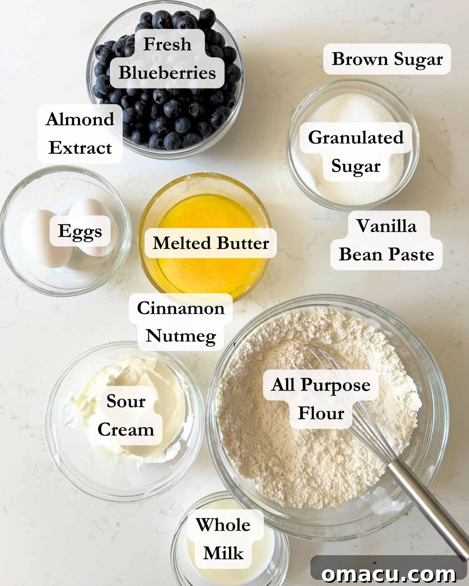 Key ingredients for blueberry sour cream coffee cake arranged in bowls on a white surface, including flour, baking powder, salt, melted butter, fresh blueberries, sugar, brown sugar, sour cream, vanilla bean paste, and almond extract.