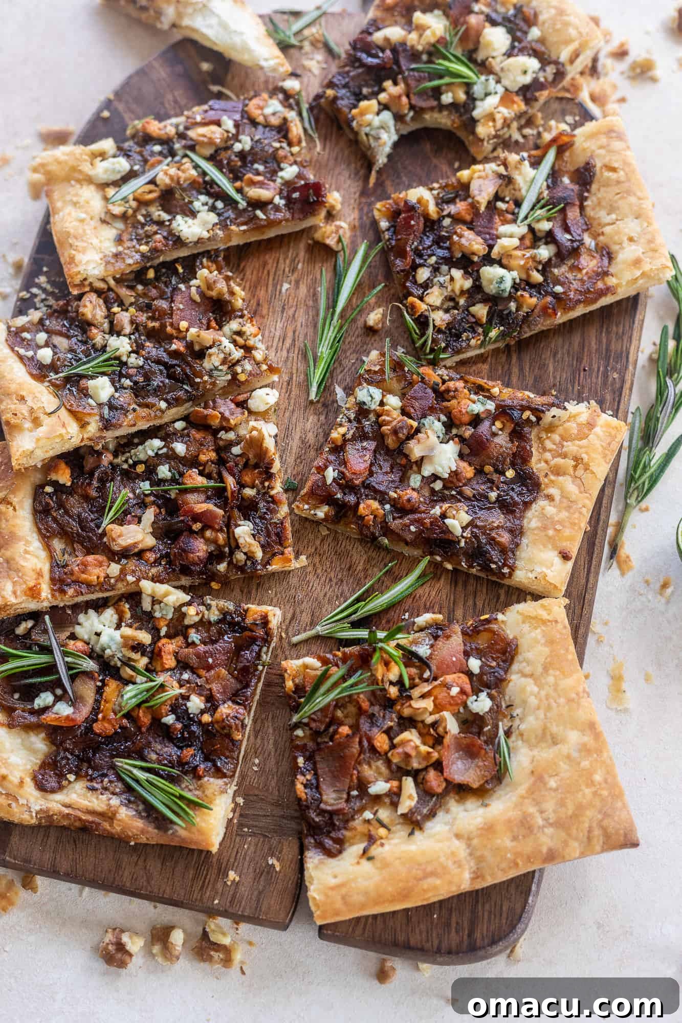 Close-up of golden-brown Caramelized Onion Puff Pastry with bacon and gorgonzola on a cutting board, garnished with fresh rosemary leaves.