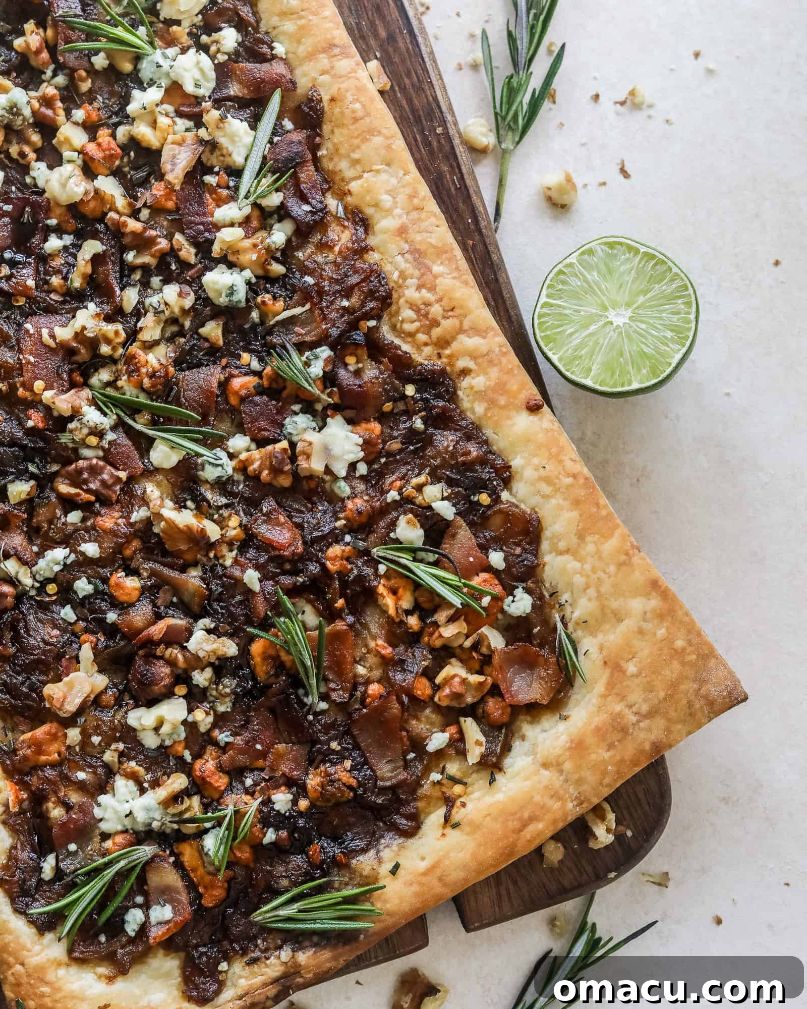 Artfully arranged Caramelized Onion Puff Pastry with bacon and gorgonzola on a wooden cutting board, with sprigs of fresh rosemary.