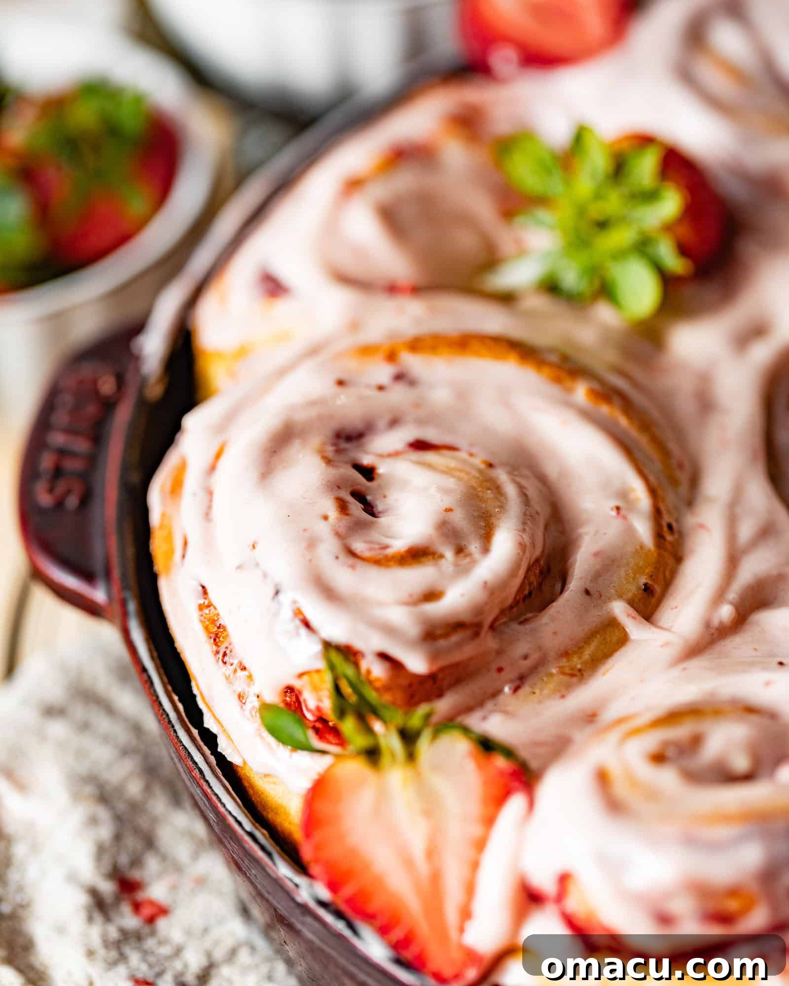 An inviting close-up of freshly baked strawberry cinnamon rolls nestled in a cast iron pan, generously adorned with strawberry glaze and slices of fresh strawberries, highlighting their gooey interior.