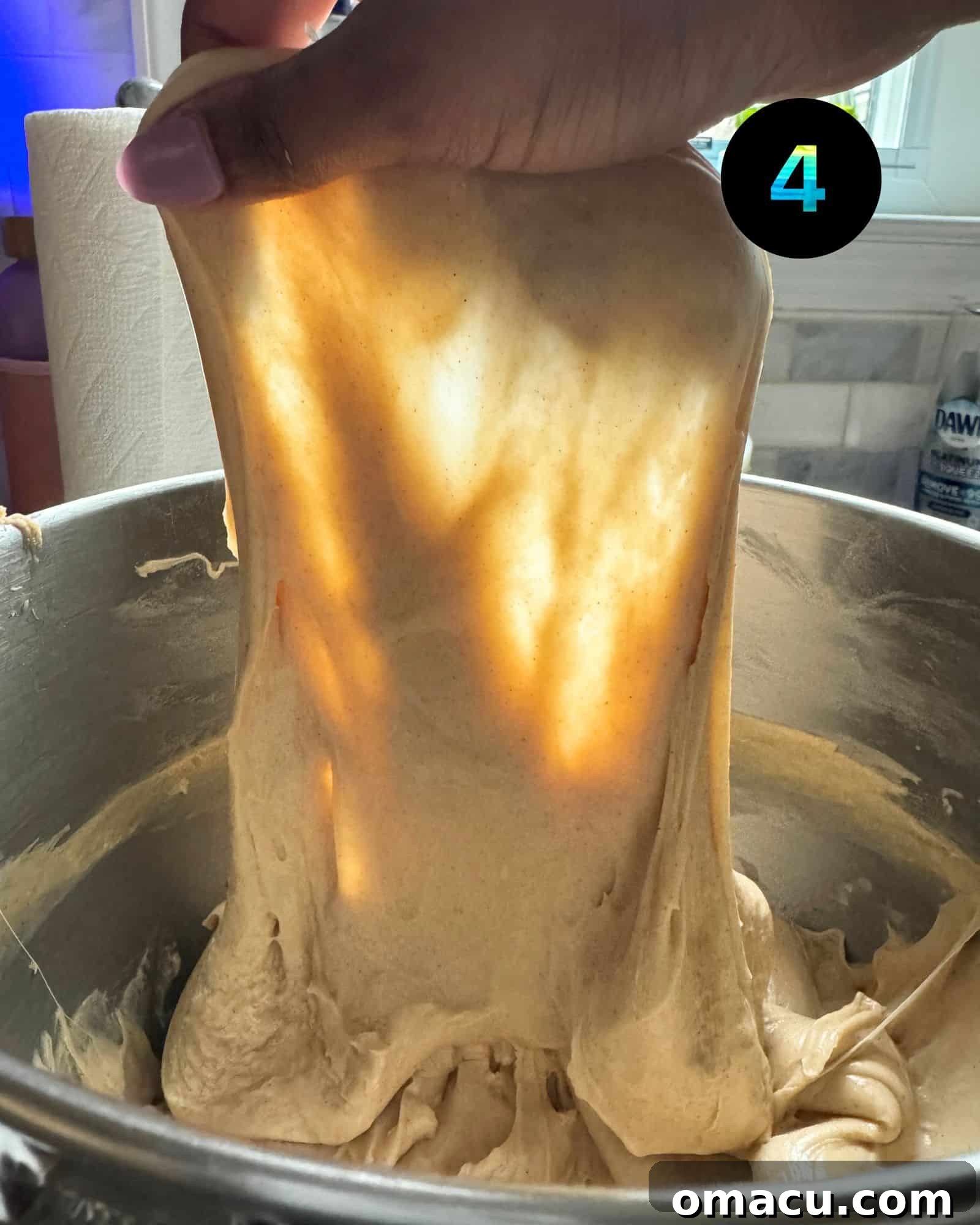 Soft, elastic cinnamon roll dough being stretched thin in a bowl to perform the windowpane test, demonstrating proper gluten development.