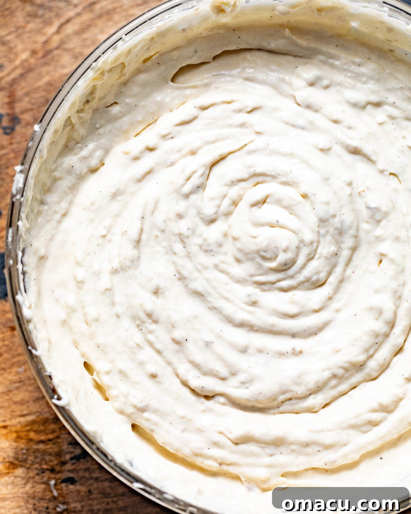 Cheesecake batter in a stand mixer bowl, now infused with mashed bananas, showing a slightly textured yet smooth consistency, ready for baking. The banana chunks are visible, promising a rich flavor.
