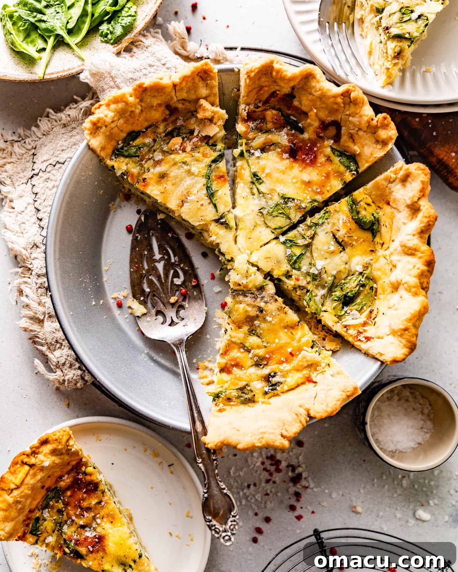 A whole Quiche Florentine in a pie dish, garnished with fresh herbs and flaky salt, ready to be served. A cake server and cheese are nearby.