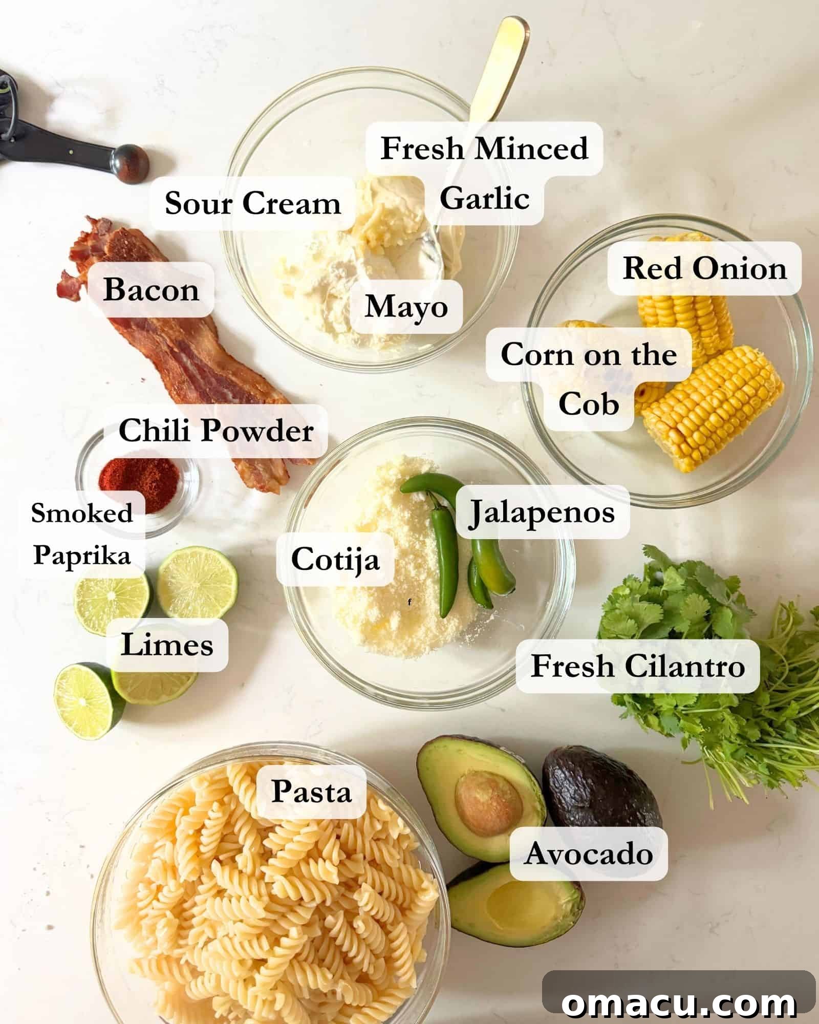 A vibrant display of ingredients arranged in bowls for making elote pasta salad: corn, jalapeños, bacon, mayonnaise, sour cream, garlic, chili powder, smoked paprika, cotija cheese, red onion, pasta, avocados, and fresh limes.