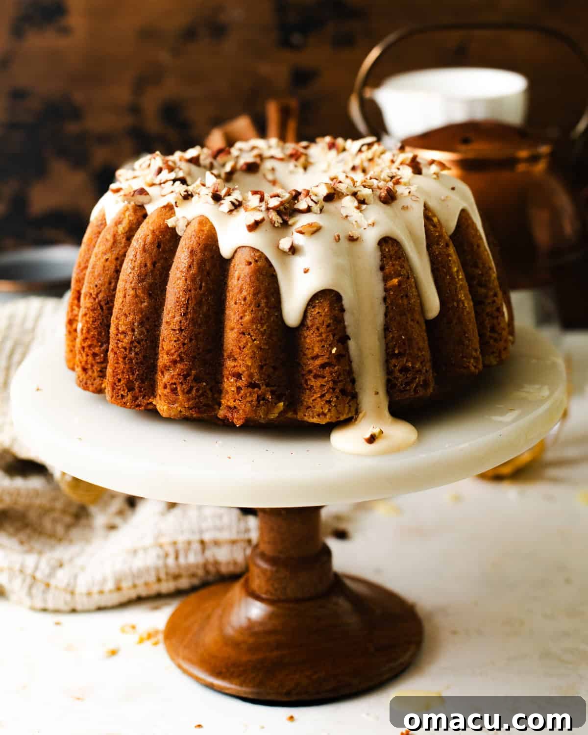 Carrot cake bundt cake on a cake stand with maple brown butter cream cheese frosting and chopped pecans on top.