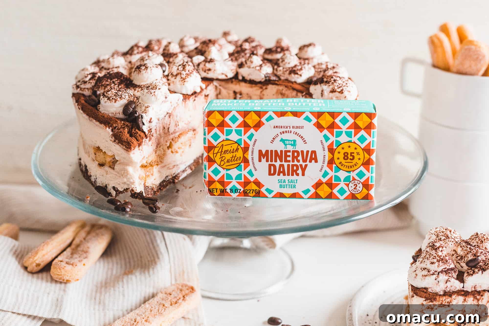 A perfectly sliced No-Bake Tiramisu Cheesecake elegantly displayed on an elevated serving dish, with an 8-ounce package of Minerva Dairy Sea Salt Butter positioned prominently in the foreground, emphasizing a key ingredient.