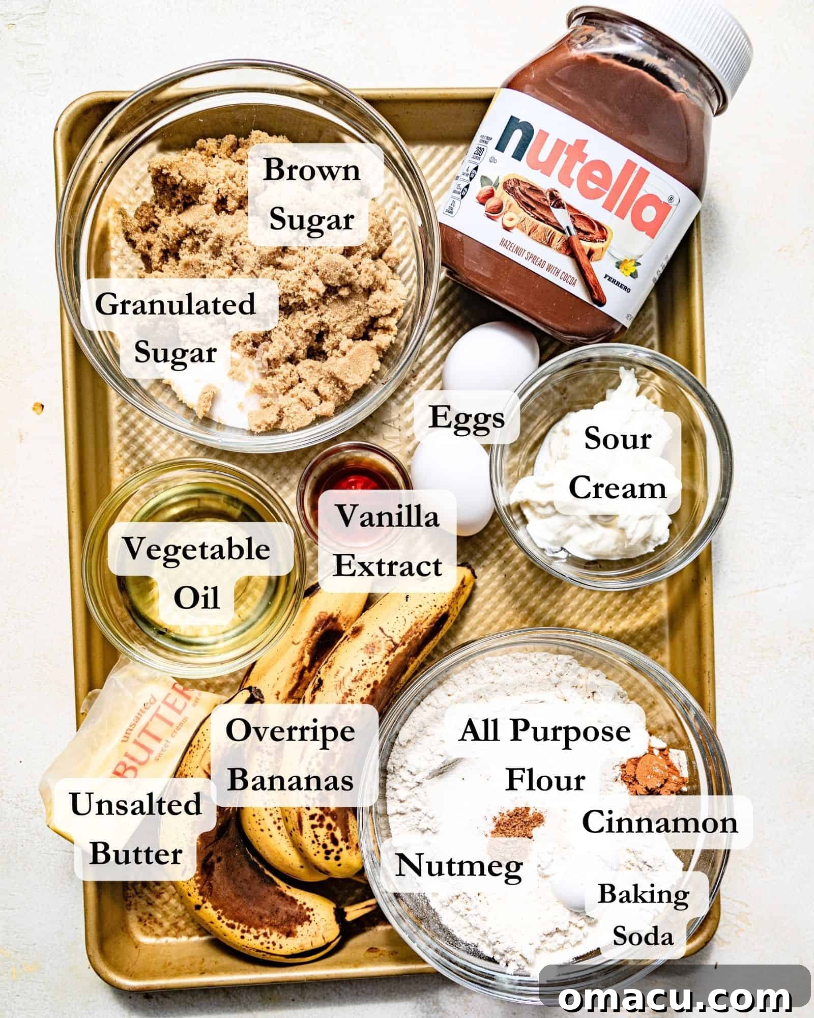 A flat lay photograph showcasing all the key ingredients needed to make banana Nutella muffins on a rustic baking sheet, including ripe bananas, a jar of Nutella, flour, sugar, brown sugar, eggs, sour cream, vegetable oil, vanilla extract, cinnamon, nutmeg, and kosher salt.