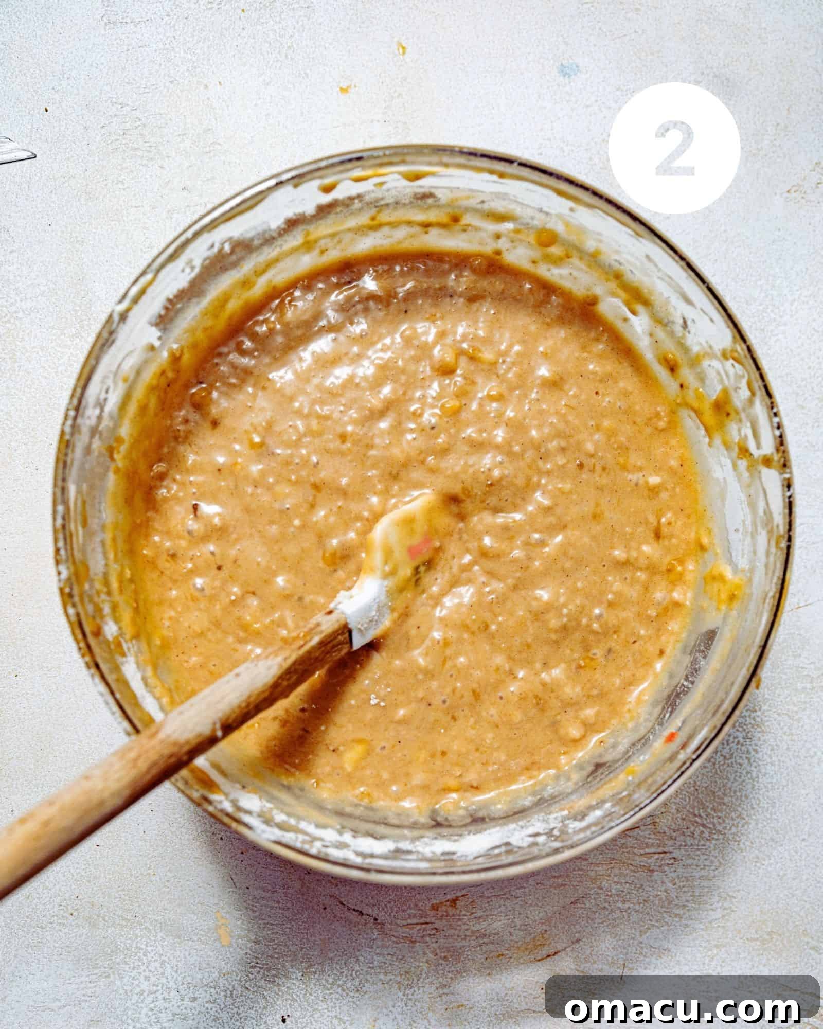 A large glass mixing bowl filled with mashed ripe bananas, partially mixed with other wet ingredients like eggs, sugars, and oil, showing a smooth yet slightly lumpy consistency.