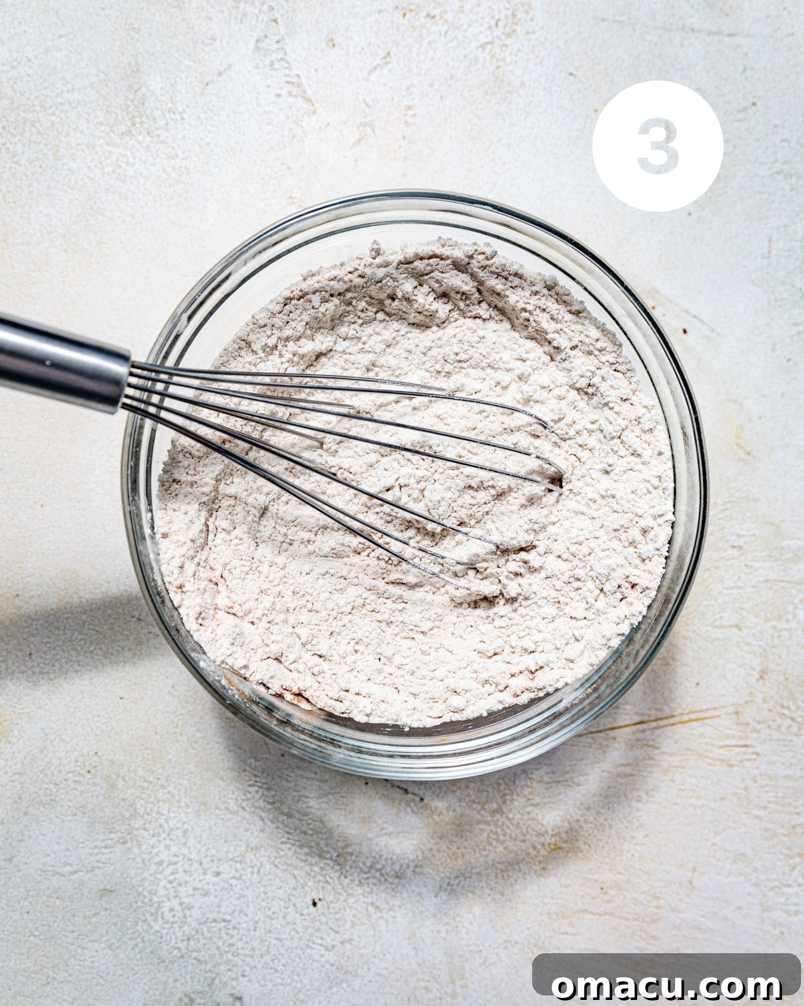 A clear glass bowl holding a mixture of dry ingredients including all-purpose flour, baking soda, kosher salt, cinnamon, and nutmeg, thoroughly whisked together.