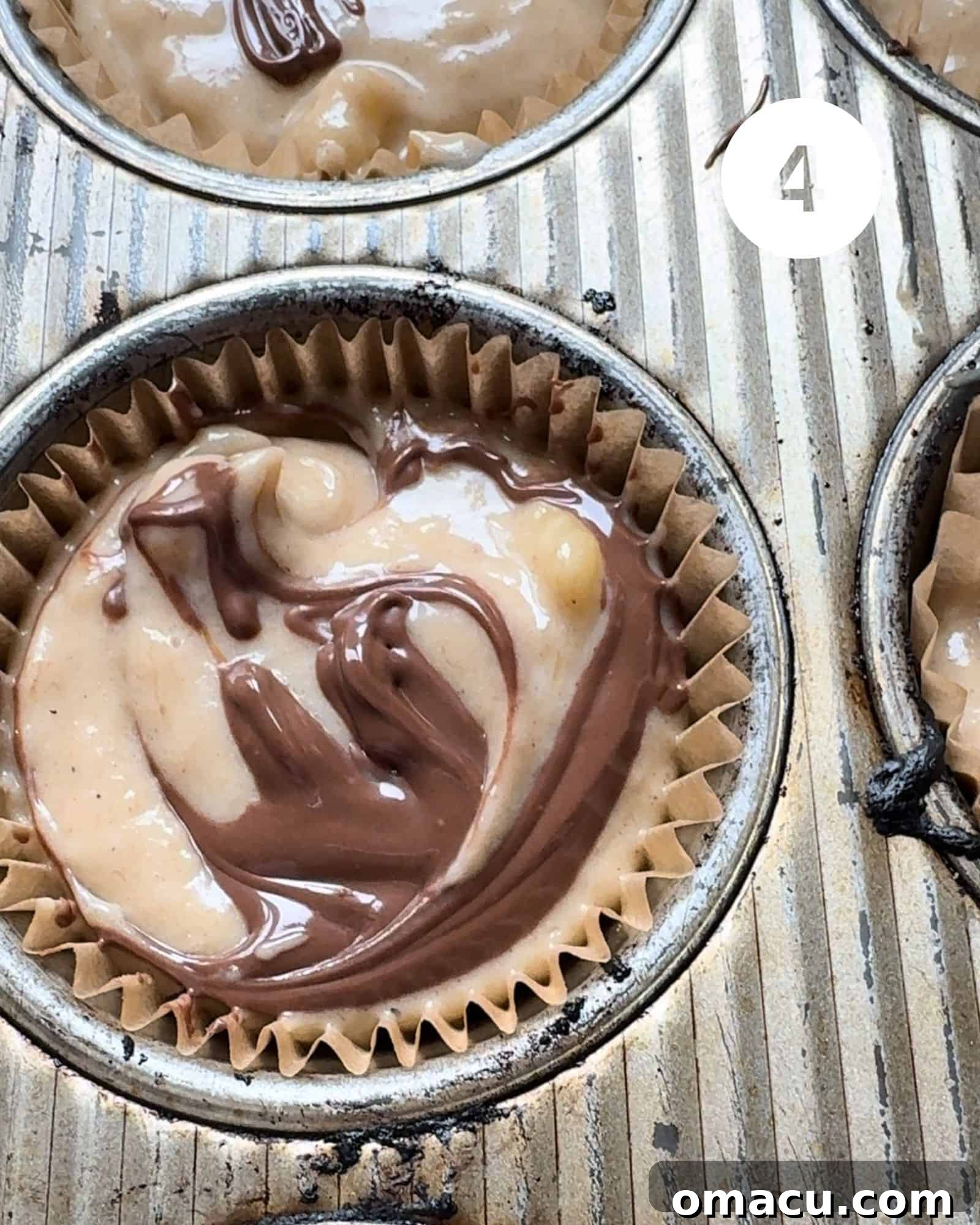 A close-up view of a muffin cup filled with batter, featuring a dollop of Nutella on top that has been gently swirled with a toothpick, creating a marble effect.