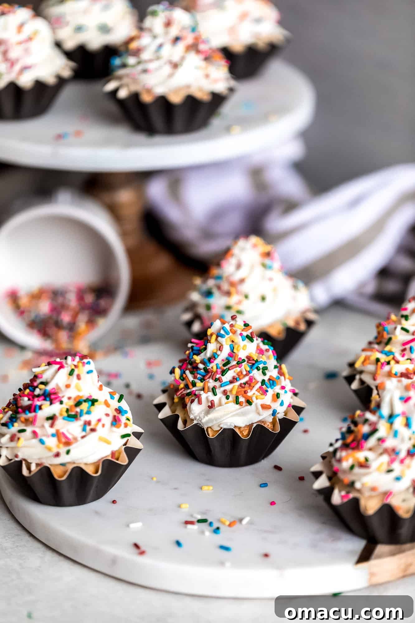 A stunning spread of Funfetti Cupcakes with Almond Buttercream on a cutting board, with more delightful cupcakes arranged on a tiered cake stand in the background, showcasing their festive appeal.