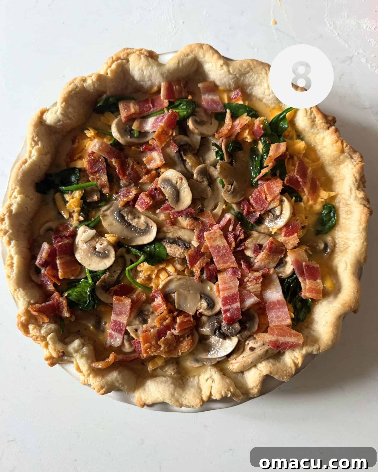 An unbaked bacon mushroom spinach quiche, fully assembled in its pie crust, layered with ingredients and ready to be placed in the oven.