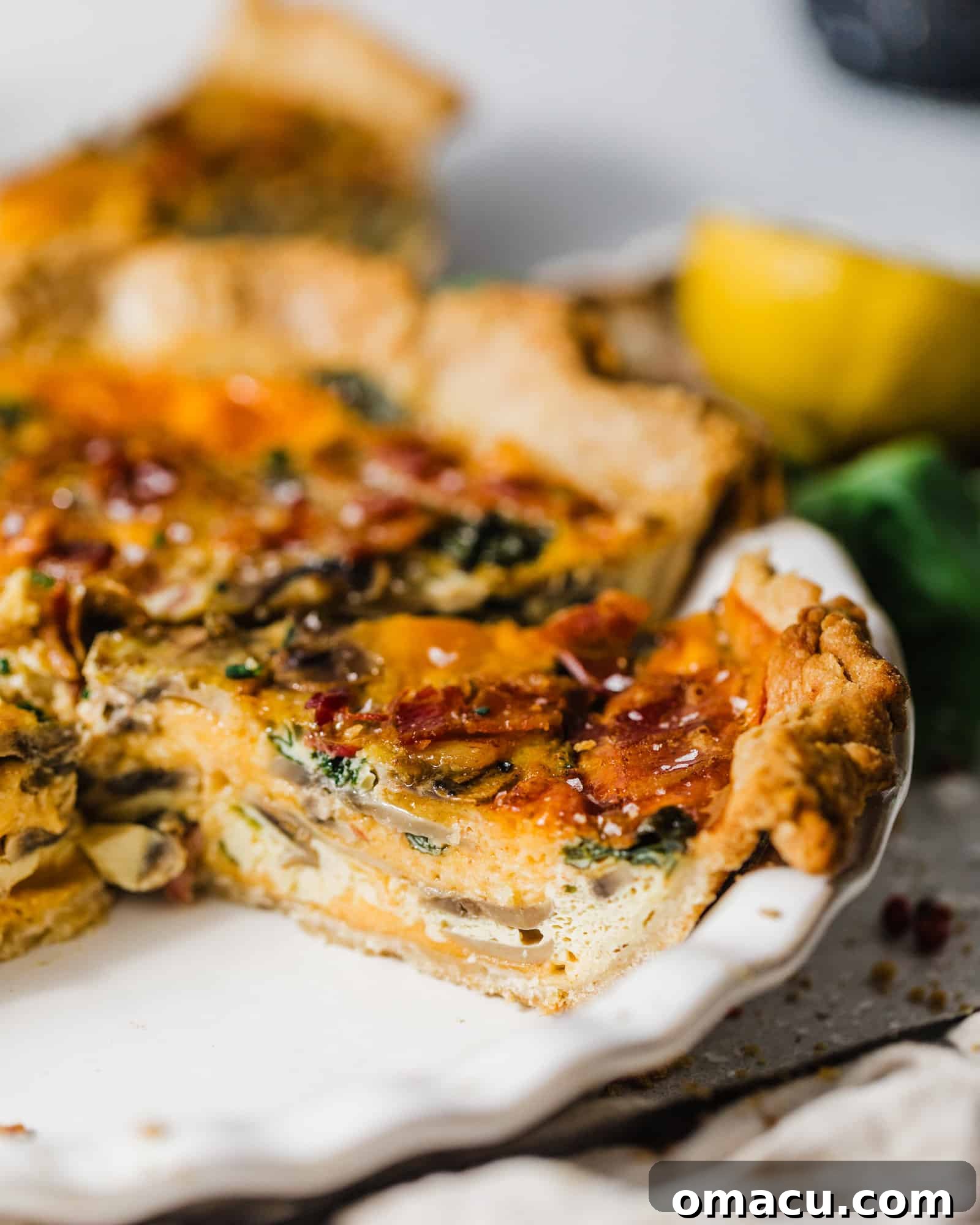 A generous slice of spinach quiche presented on a plate, with a portion already removed, revealing the rich layers of bacon, mushrooms, and cheese within the creamy filling.
