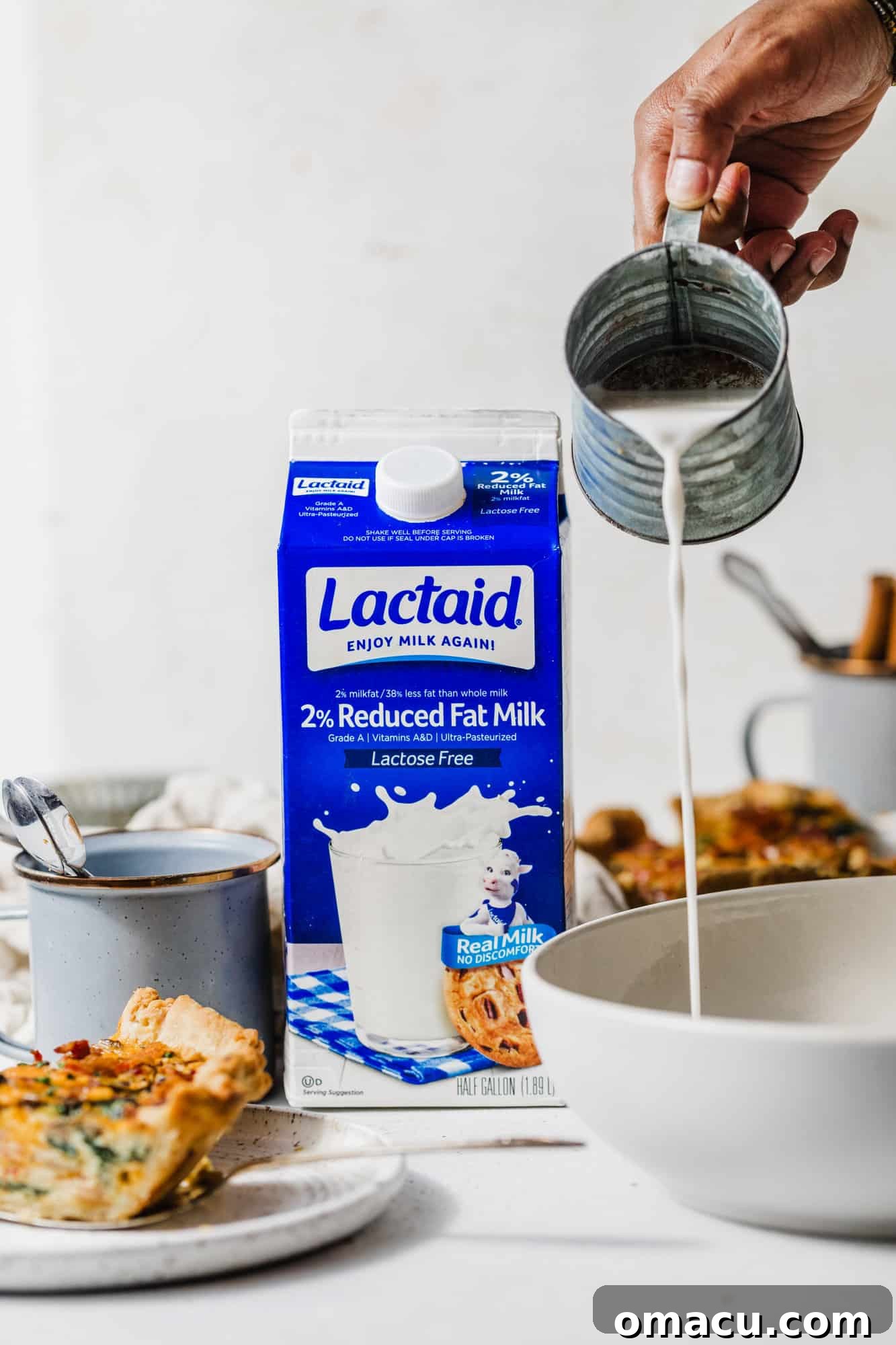 A carton of fresh milk sits beside a large mixing bowl, with a hand gently pouring milk into the bowl, highlighting a key ingredient for the quiche custard.