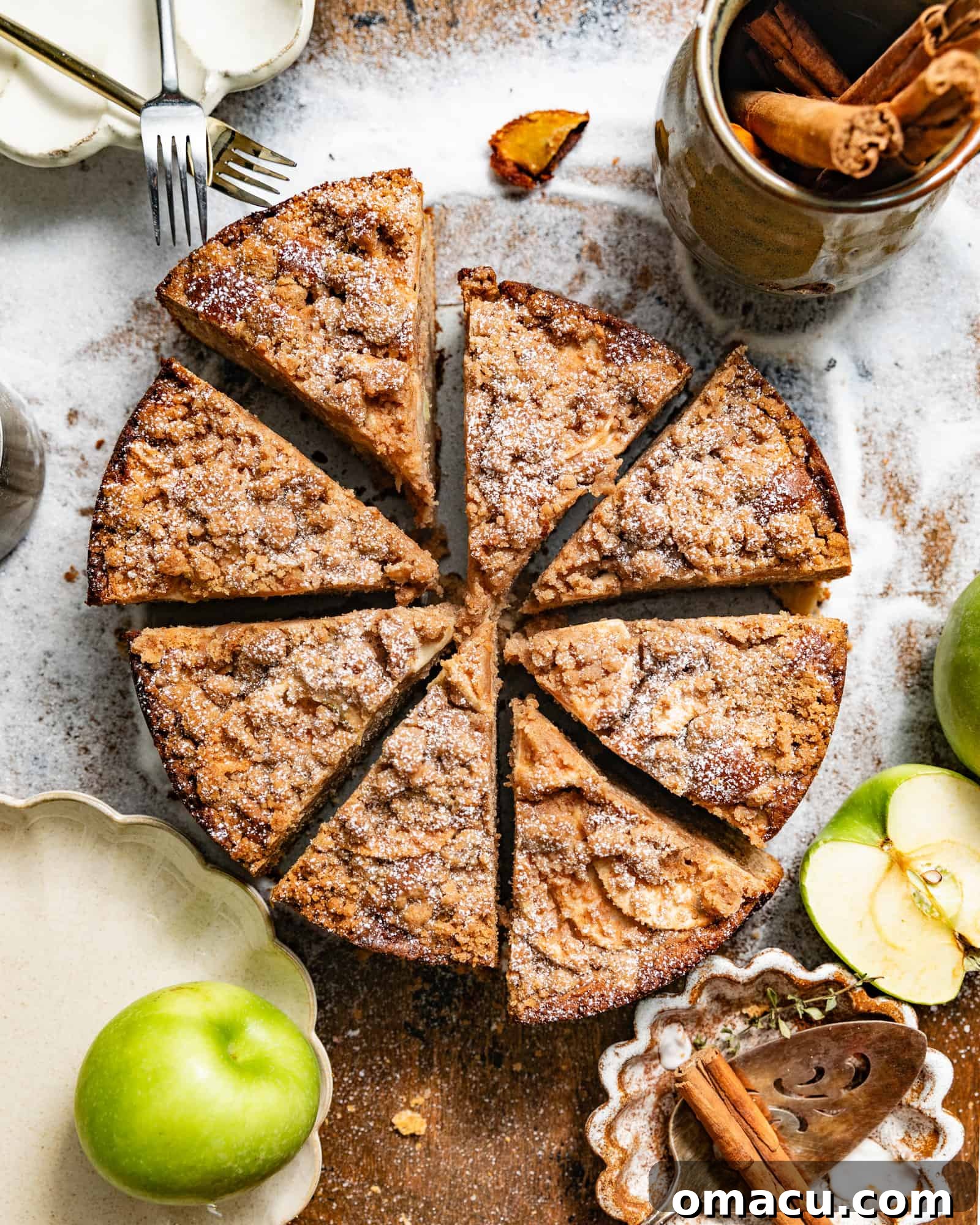 Delicious Dutch Apple Cake with a rich crumb topping, served on a rustic brown surface alongside fresh apple slices, a ceramic mug, and cinnamon sticks.