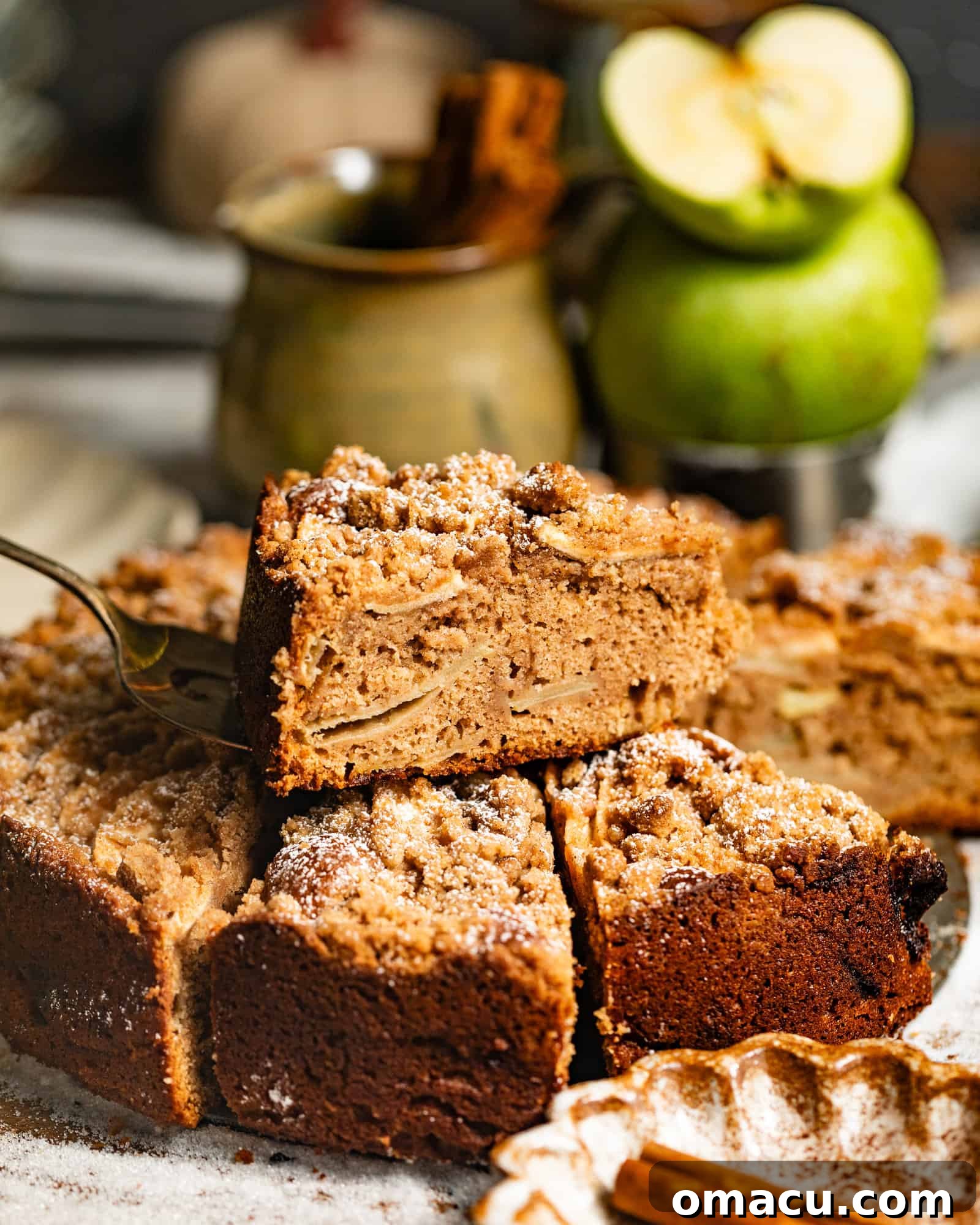 A slice of moist Dutch Apple Cake topped with streusel, showing the tender apple chunks within, presented on a white plate with another slice resting on top.