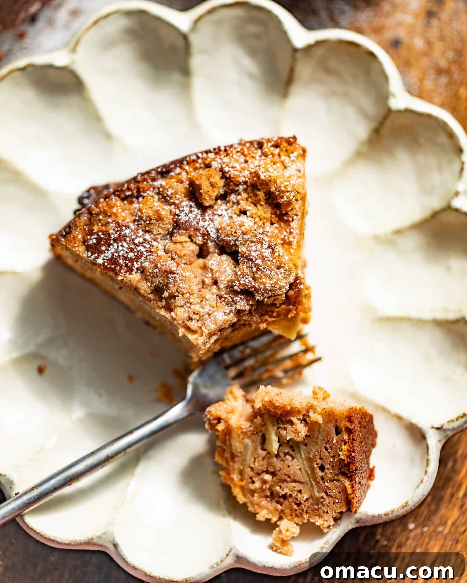 A perfectly baked slice of Dutch Apple Cake on a white plate with a fork, ready to be enjoyed, highlighting its moist texture and crumb topping.