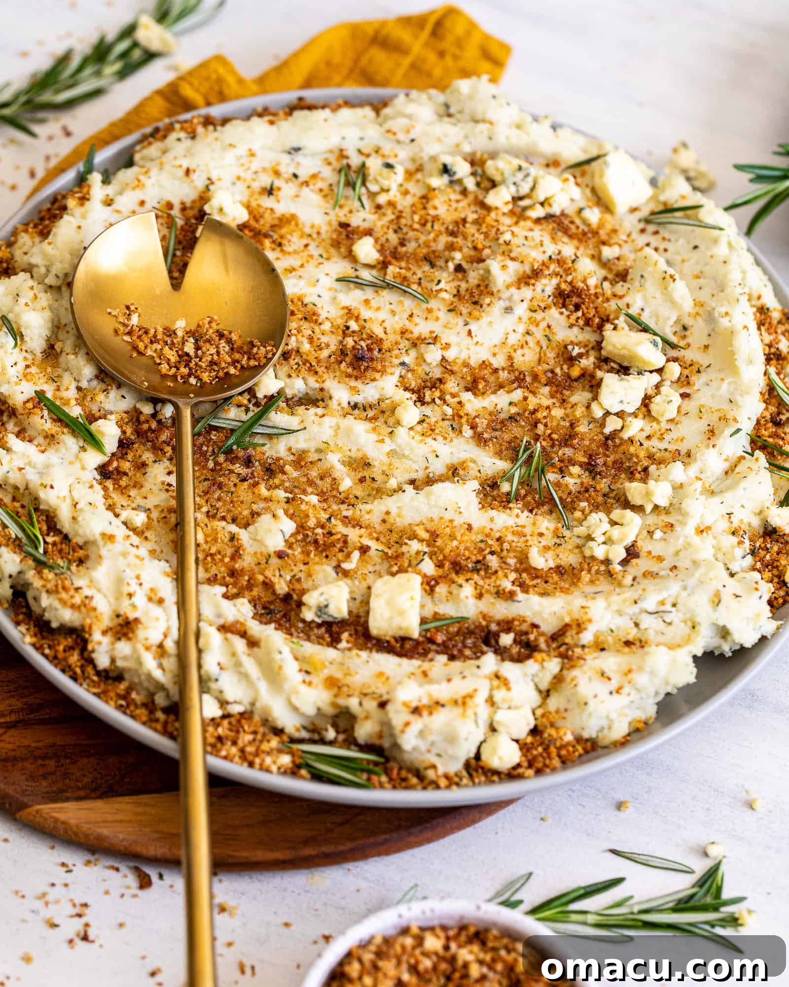 A gold spoon placed over a large bowl of Blue Cheese Mashed Potatoes with Toasted Breadcrumbs, ready to be served.