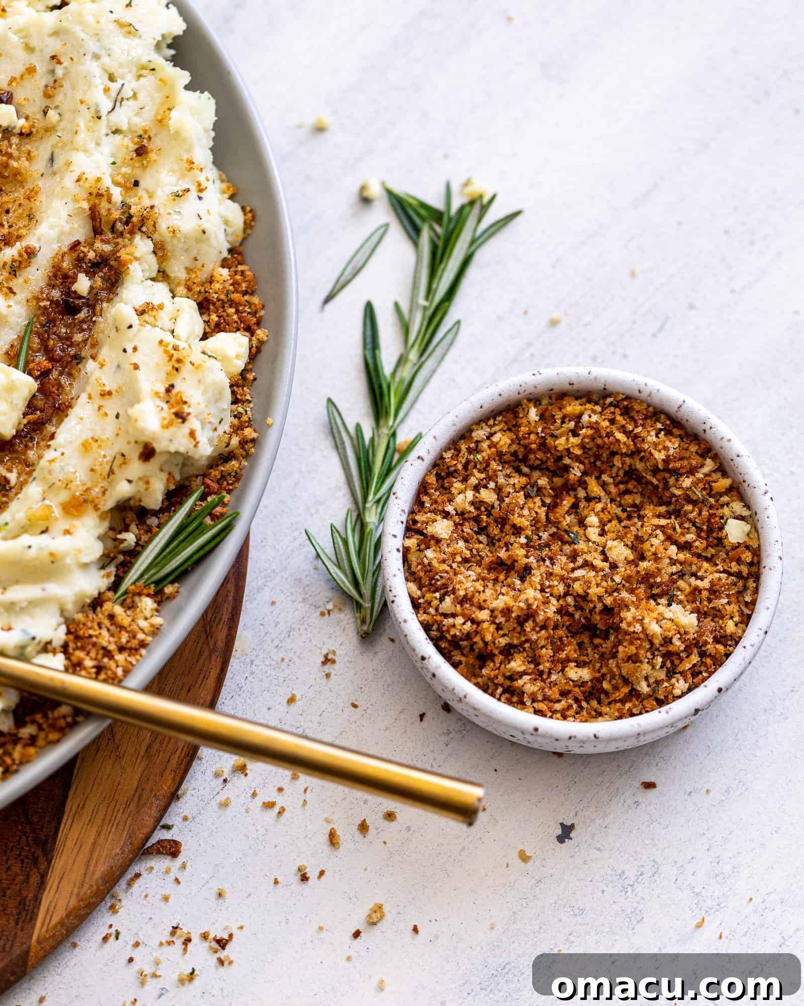 A small bowl of golden toasted breadcrumbs sits beside a serving of creamy mashed potatoes, garnished with a sprig of fresh rosemary.
