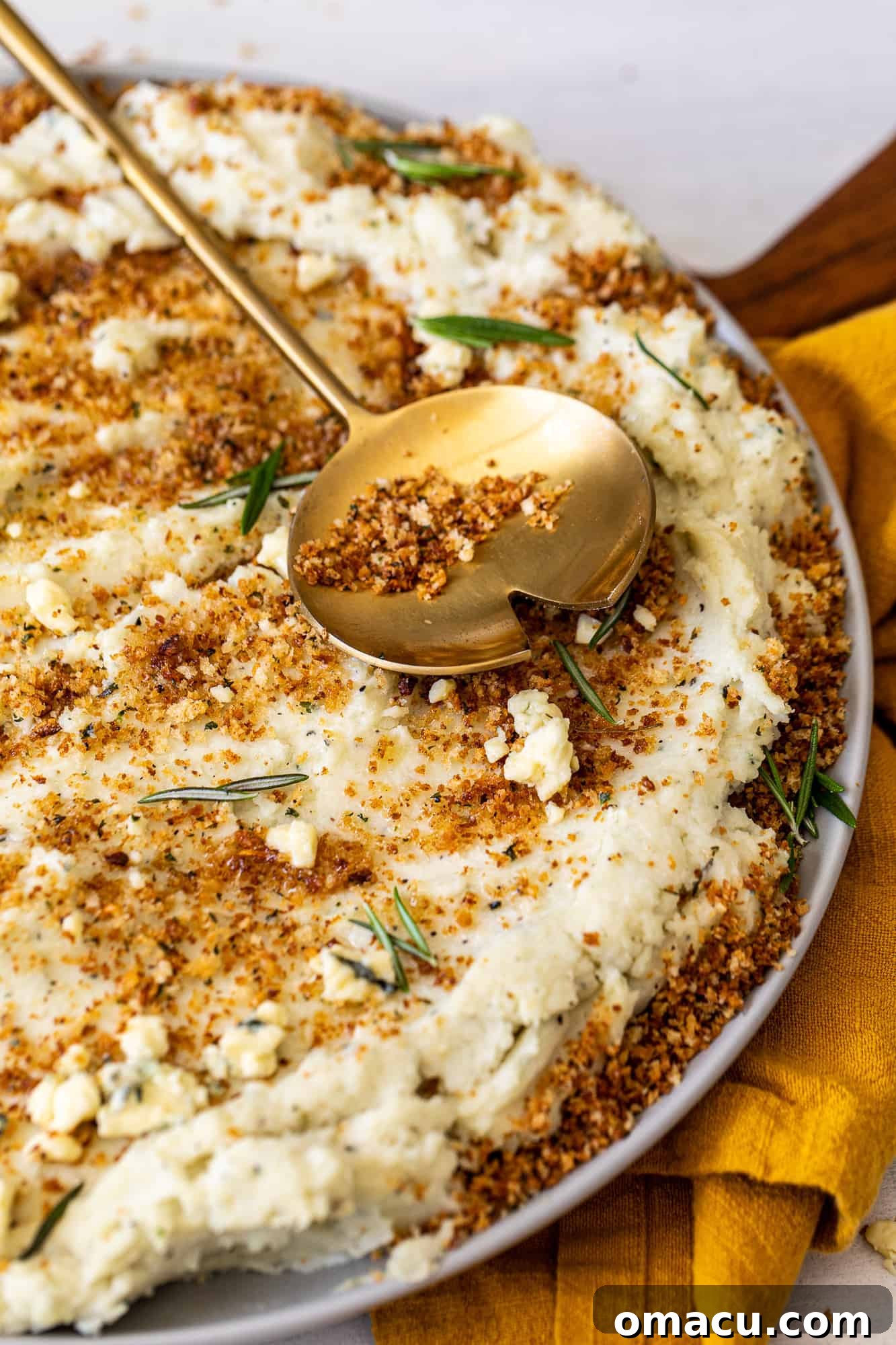 A gold spoon is carefully placed over a large bowl of Blue Cheese Mashed Potatoes with Toasted Breadcrumbs, emphasizing its inviting texture.