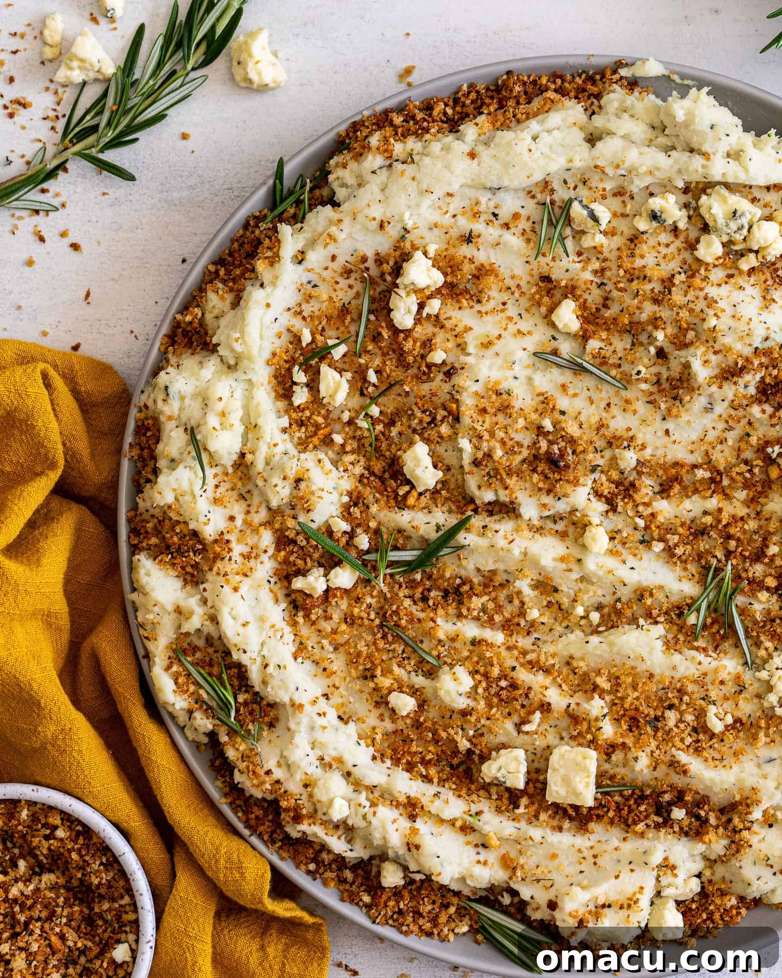 A large bowl of mashed potatoes is presented alongside a mustard-colored cloth and a small bowl filled with golden toasted breadcrumbs.